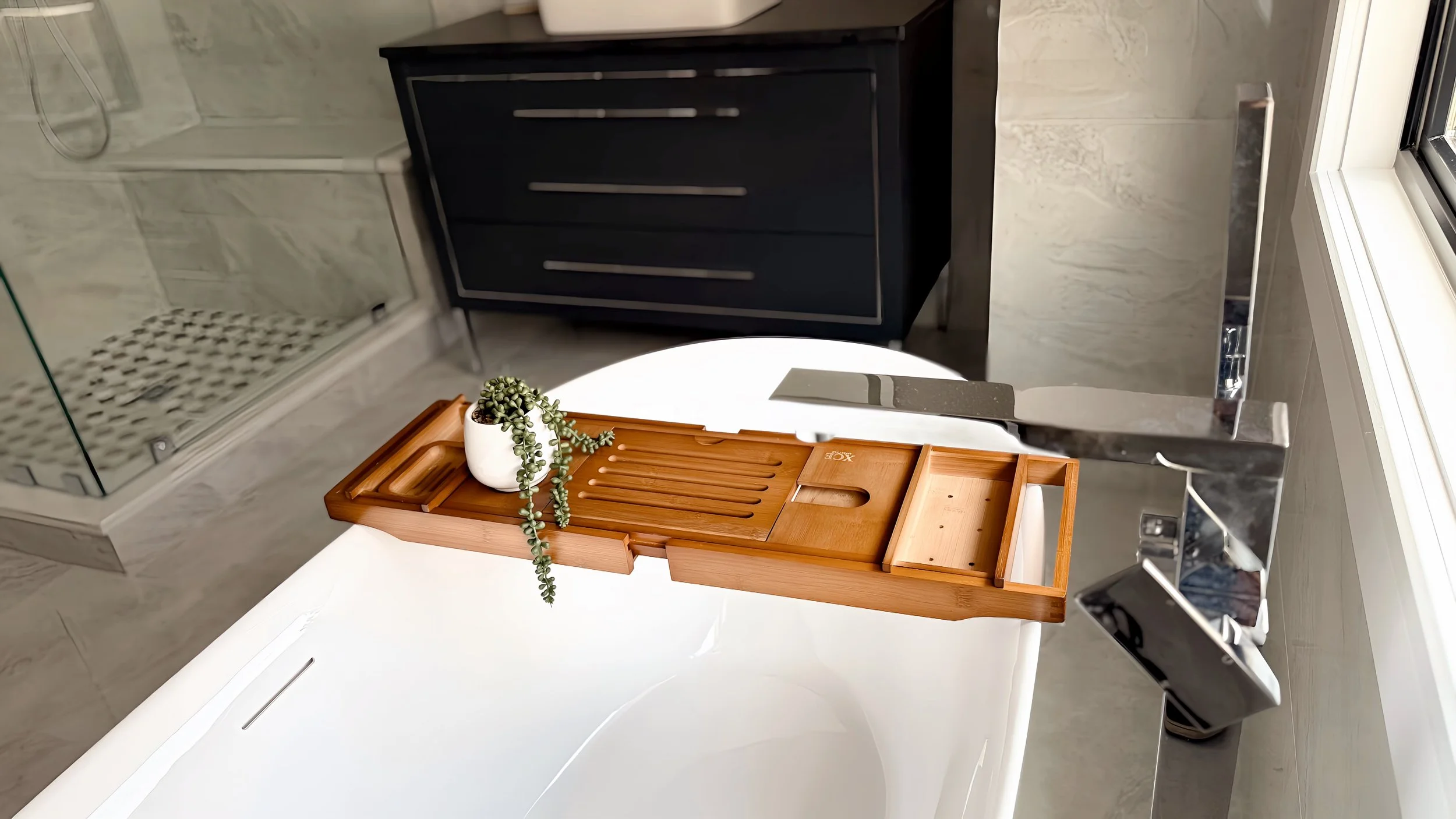 Bathroom decor with a small potted succulent plant on a wooden tray placed on a white surface, with a black cabinet and shower tiles visible in the background.