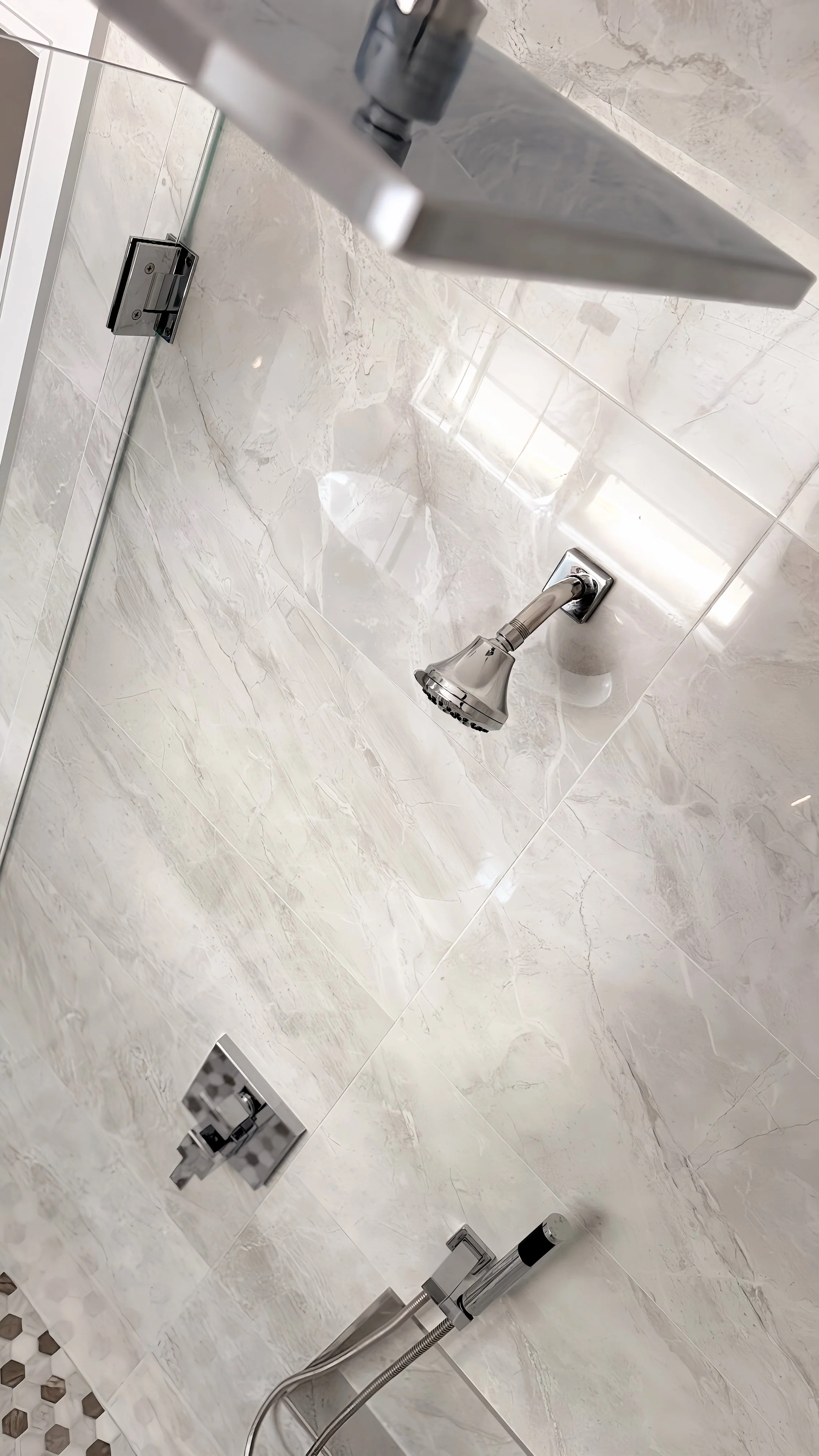 Close-up of a shower area with a handheld showerhead, wall-mounted shower controls, and a glass partition with beige marble tiles.