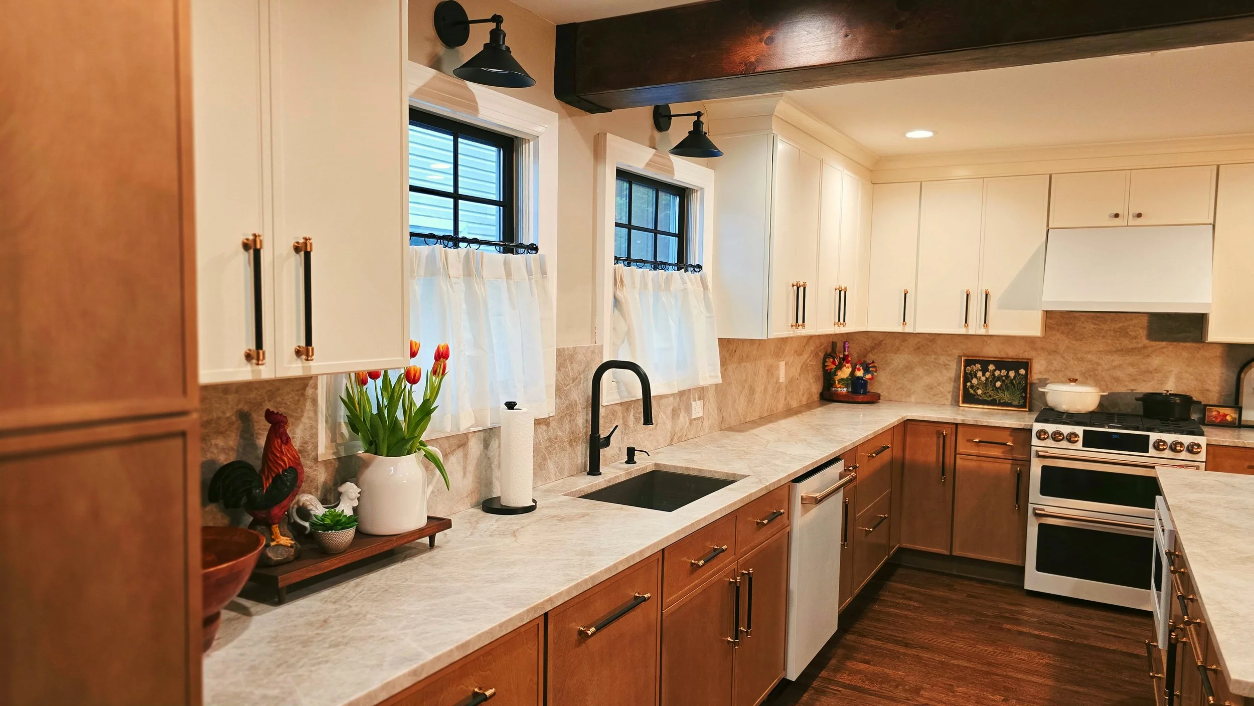 A kitchen with white upper cabinets, brown lower cabinets, a white marble countertop, a black sink with a black faucet, two windows with white curtains, and some decorative items including tulips and a rooster figurine.