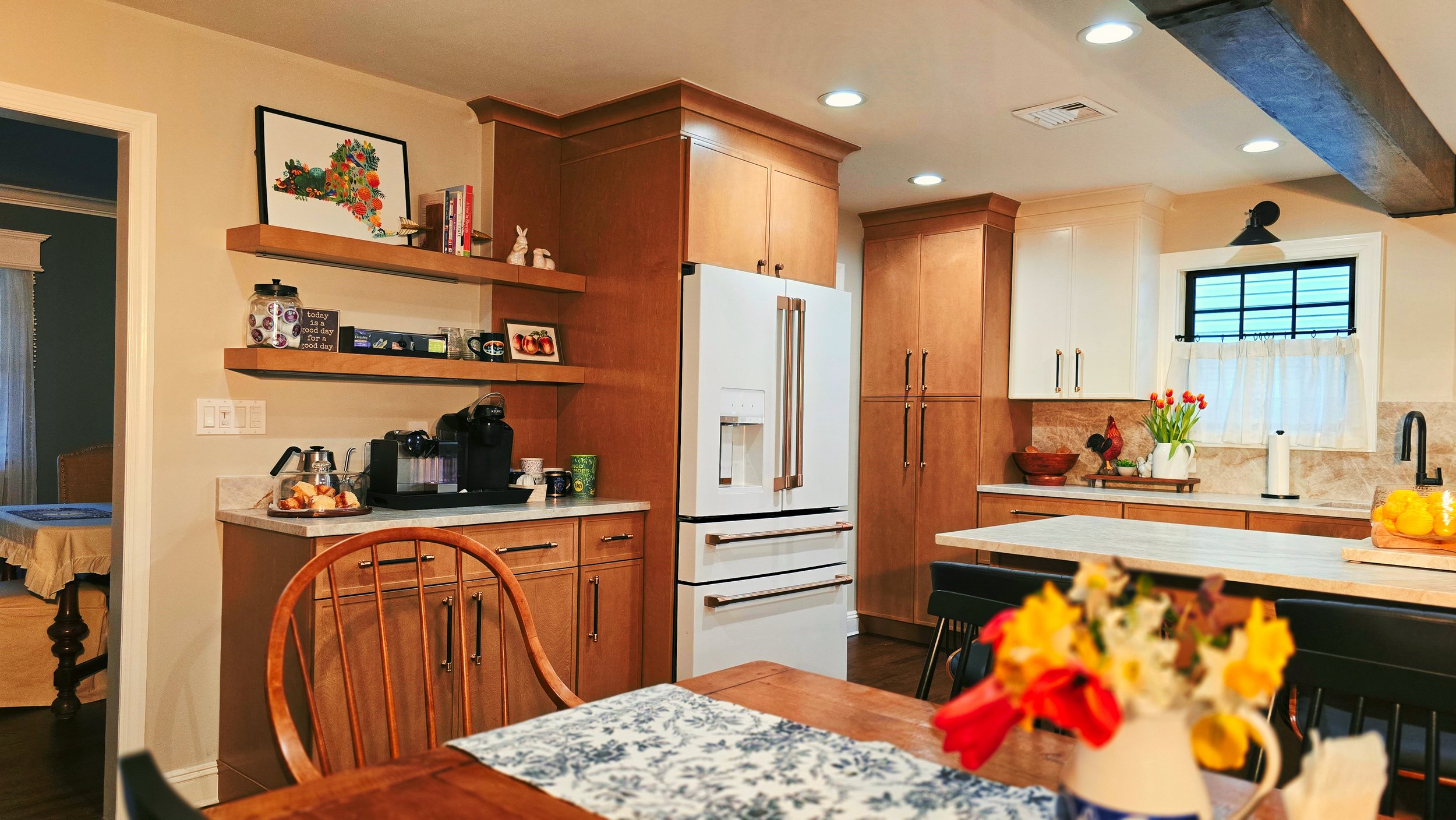 A kitchen with wooden cabinets, a white refrigerator, a window with curtains, a coffee station, and a dining table with a floral centerpiece.