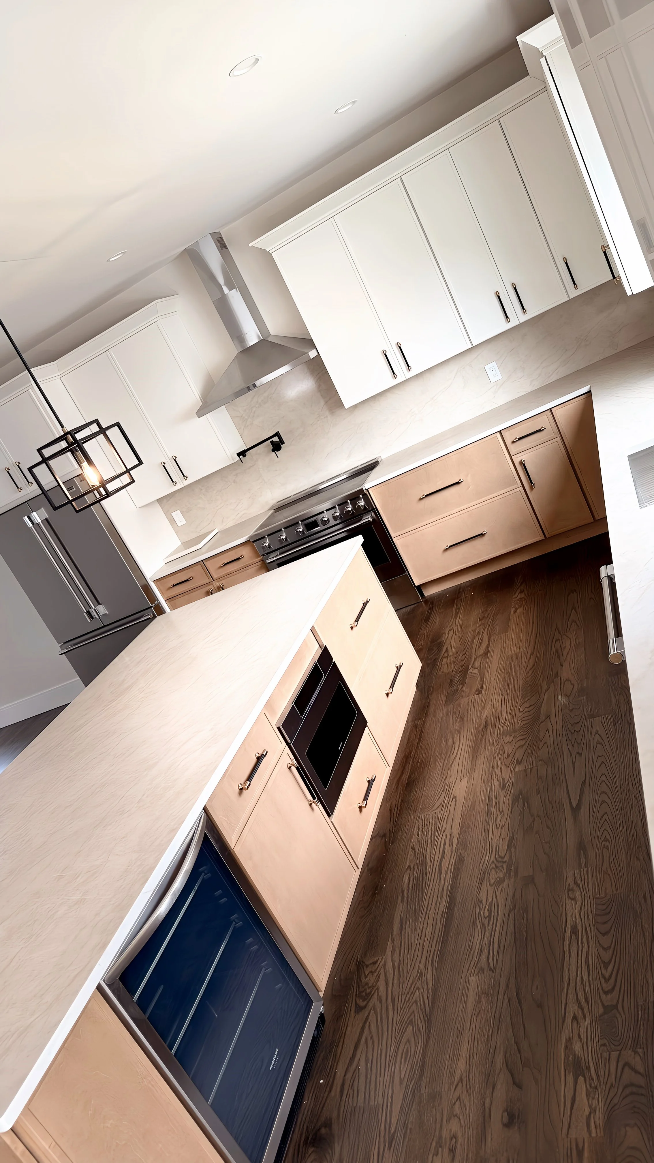 A modern kitchen with white upper cabinets, light wood lower cabinets, a large island, and dark wood flooring. Includes stainless steel appliances and a geometric chandelier.