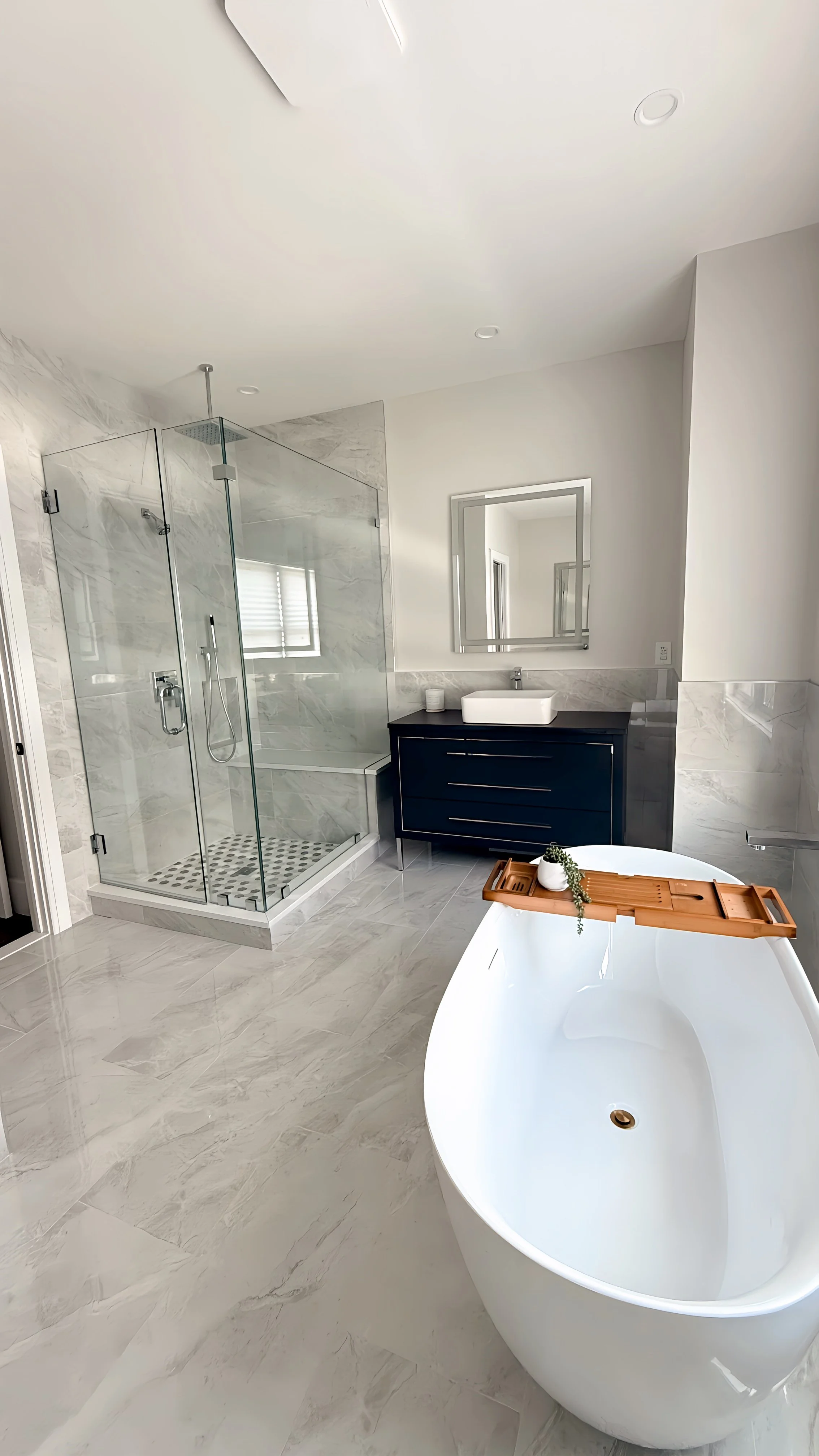 Modern bathroom with a white freestanding bathtub, a glass-enclosed shower with rainfall and handheld showerheads, a black vanity with a rectangular sink, and a mirror above. The room features gray marble tiles and a minimalist aesthetic.
