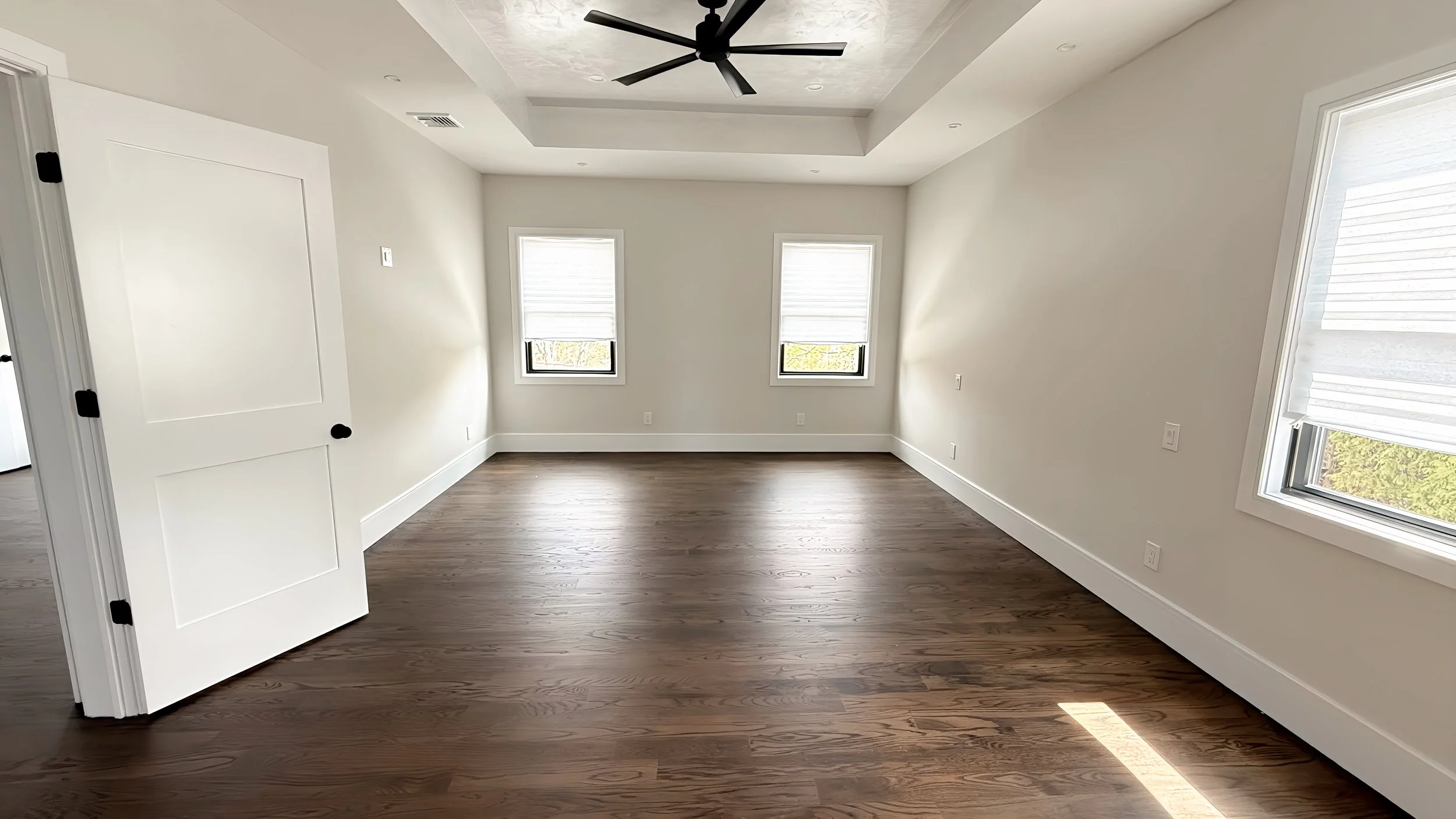 Empty room with three windows, white walls, wood flooring, ceiling fan, and a door partially open.