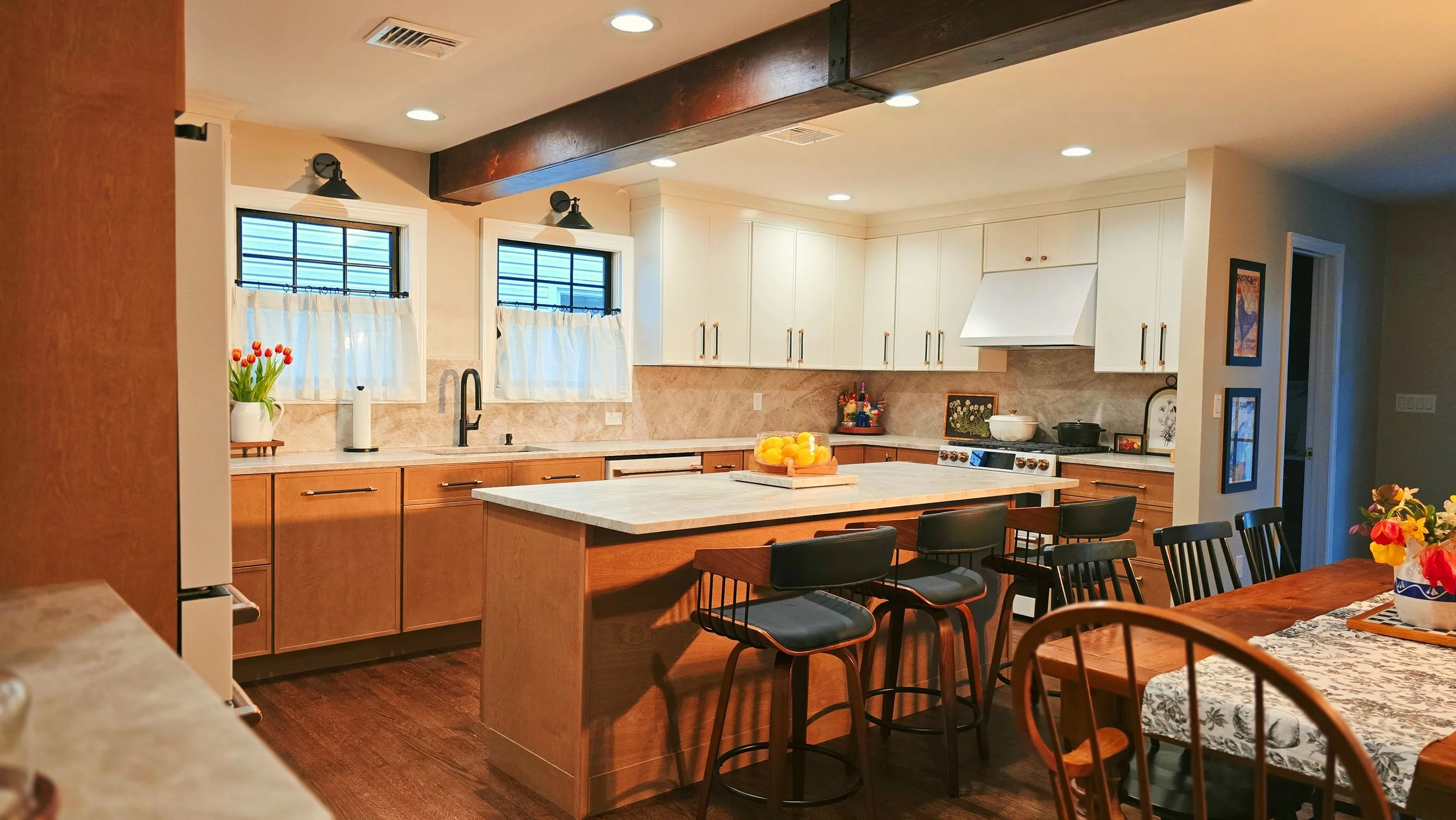A spacious kitchen with wooden cabinets, a large island with a white countertop, and a dining area with chairs. There are two windows with white curtains, a vase of red tulips, and a bowl of lemons on the island. Black chairs surround the dining table, and there is a floral tablecloth and additional floral decor on the side.