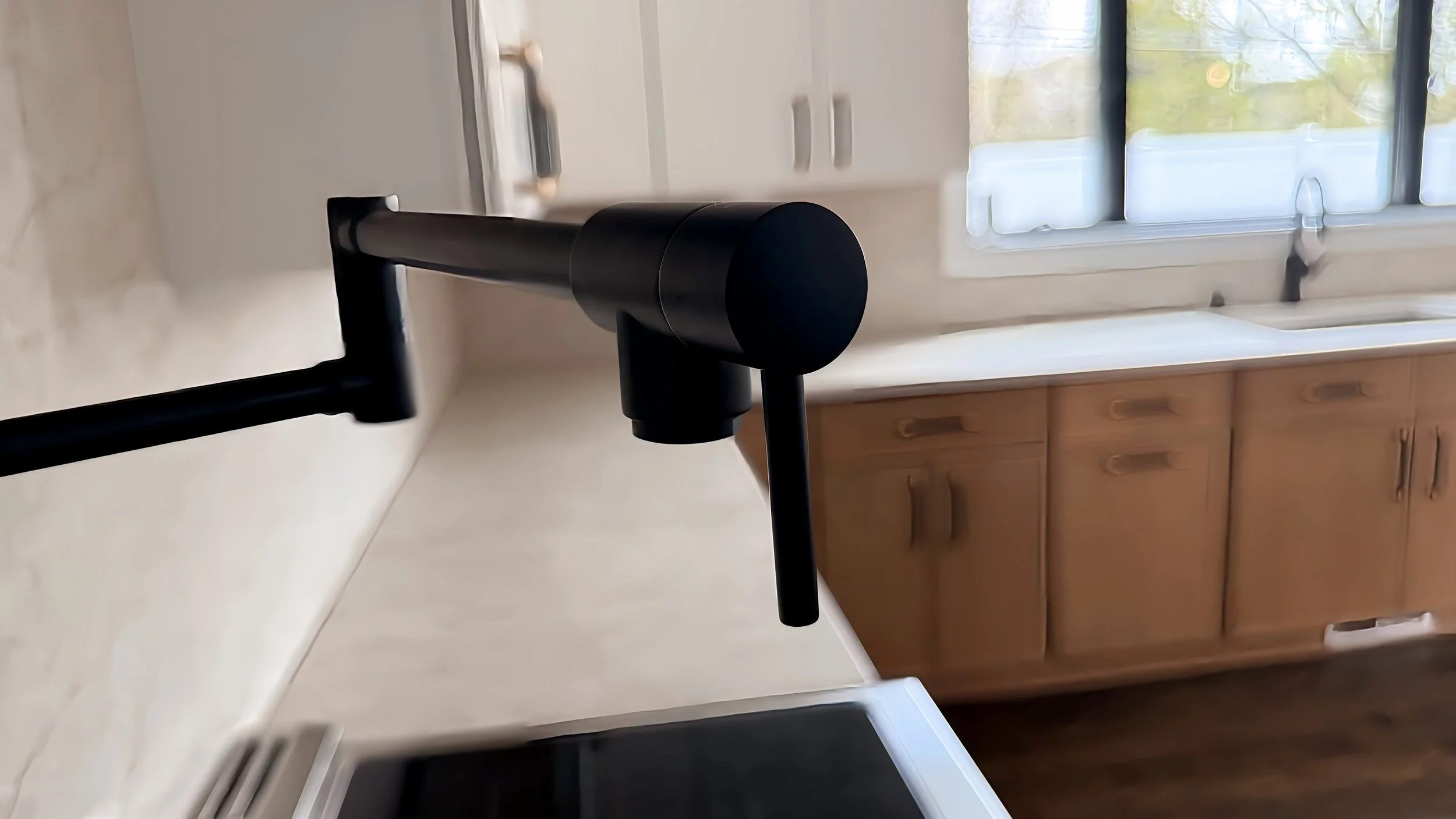 Black kitchen faucet with curved spout on a white kitchen counter near a window with blinds, wooden cabinets below, and outdoor scenery visible through the window.