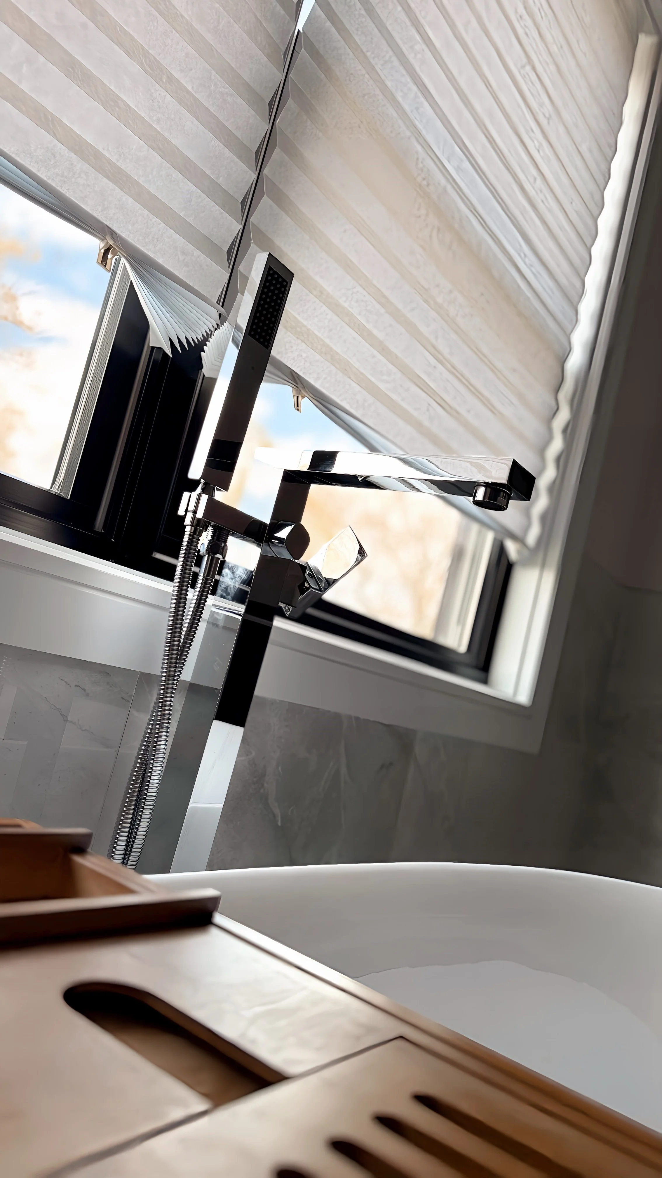 Close-up view of a modern bathroom with a freestanding bathtub, a window with blinds, and a sleek chrome-finished showerhead.
