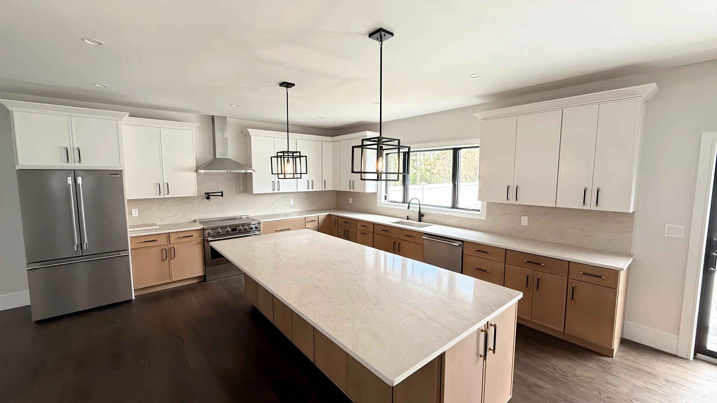 Modern kitchen with white upper cabinets, wood lower cabinets, a large marble island, stainless steel appliances including a refrigerator, oven, and range hood, pendant lighting fixtures, and a window overlooking the outside.