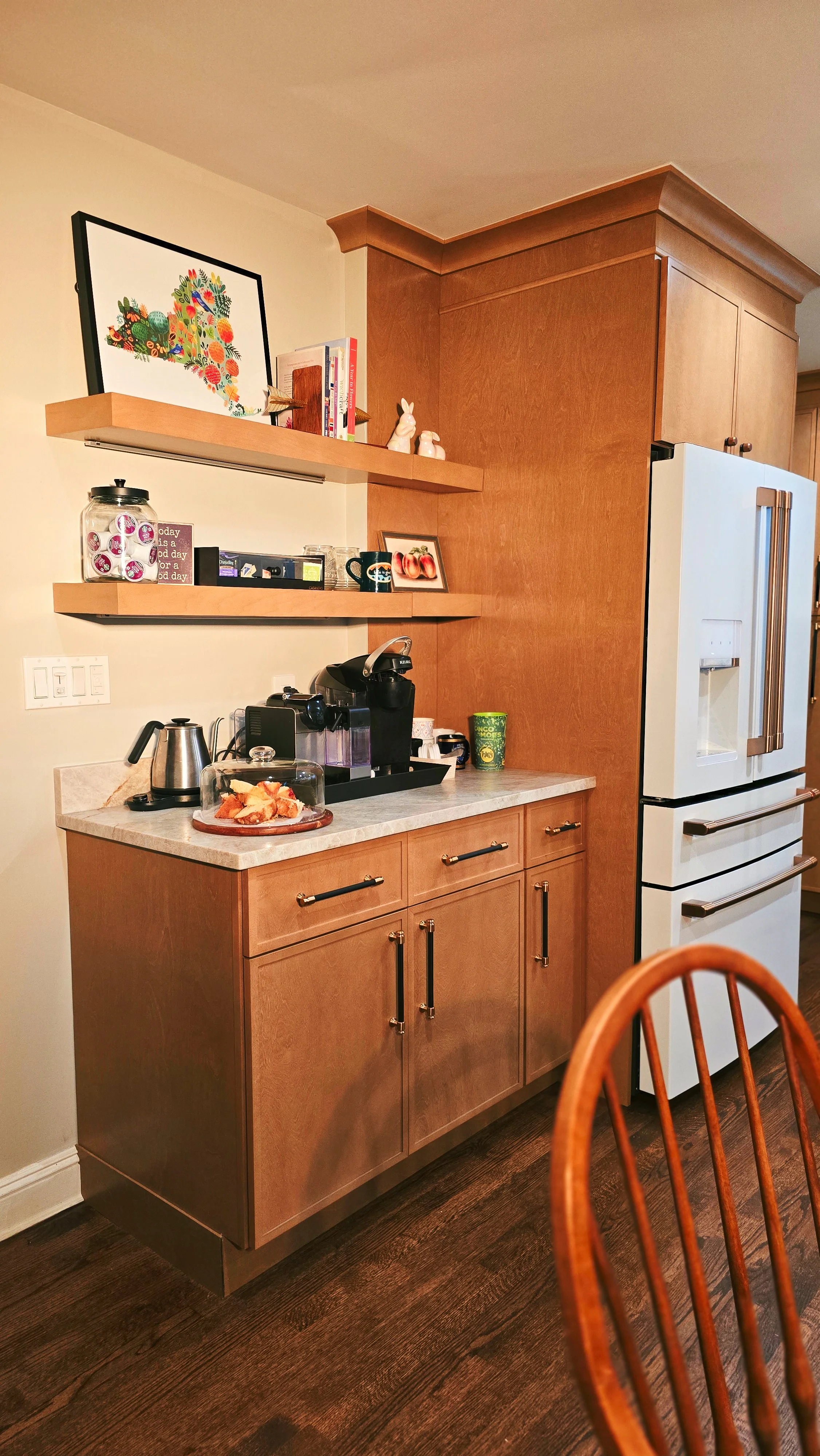 A kitchen corner with wood cabinets, a white refrigerator, and a beige countertop. On the countertop, there is a coffee machine, a kettle, and a plate of cookies. Two floating wooden shelves hold books, decorative rabbits, and framed pictures. A wood