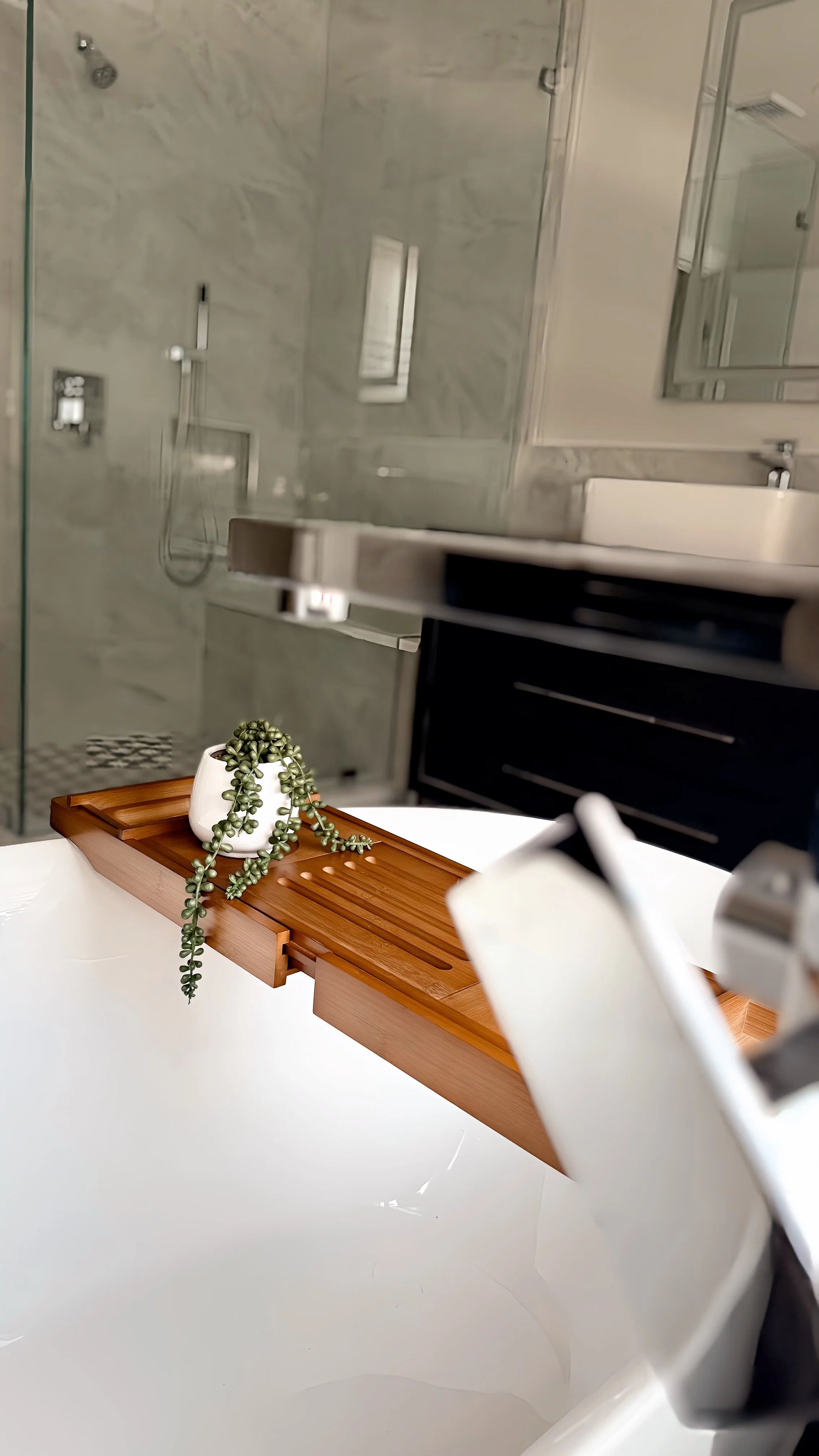 Close-up of a bathroom with a shower area, a wooden tray on a white surface, and a small potted plant with hanging green leaves.