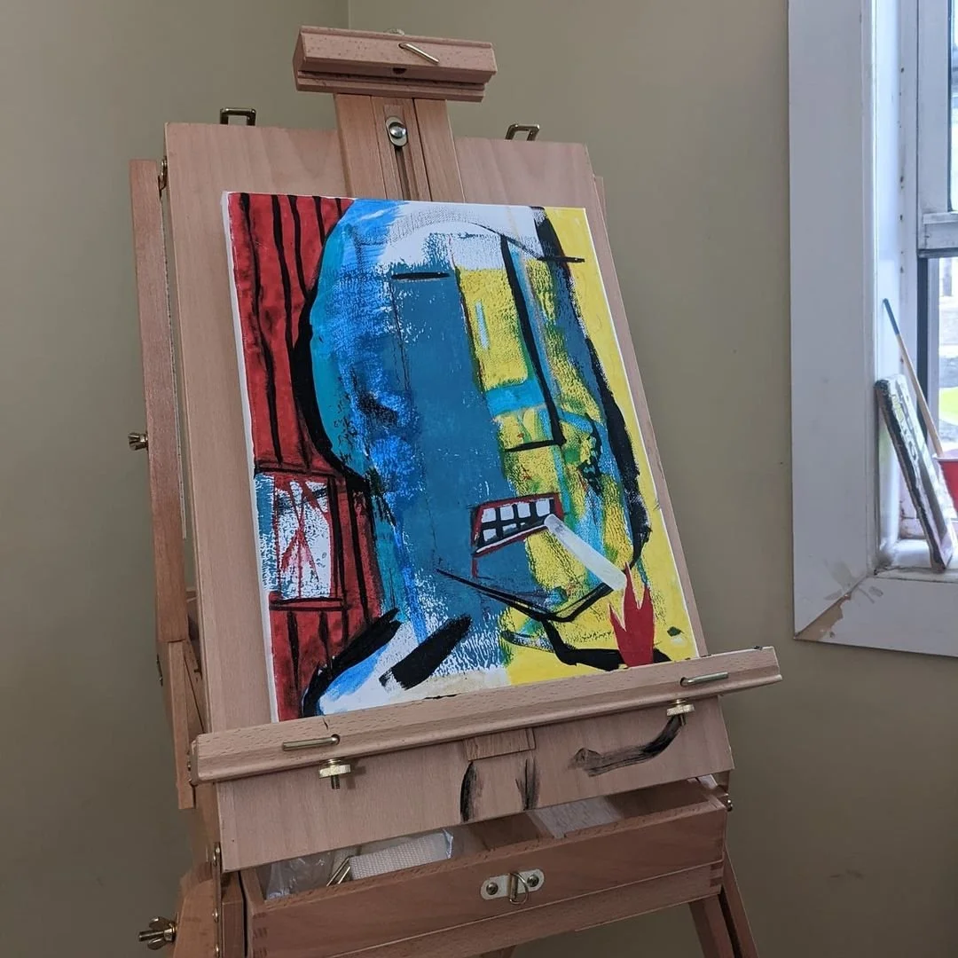 An abstract landscape painting on an easel featuring bold colors like blue, yellow, red, and black with expressive brushstrokes, positioned near a window in a room.