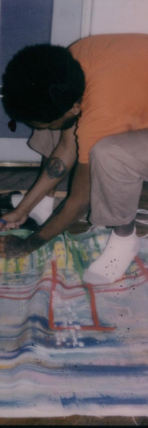 Person with black hair and tattoos wearing an orange shirt and beige pants, sitting on a couch, reaching down on a colorful rug.