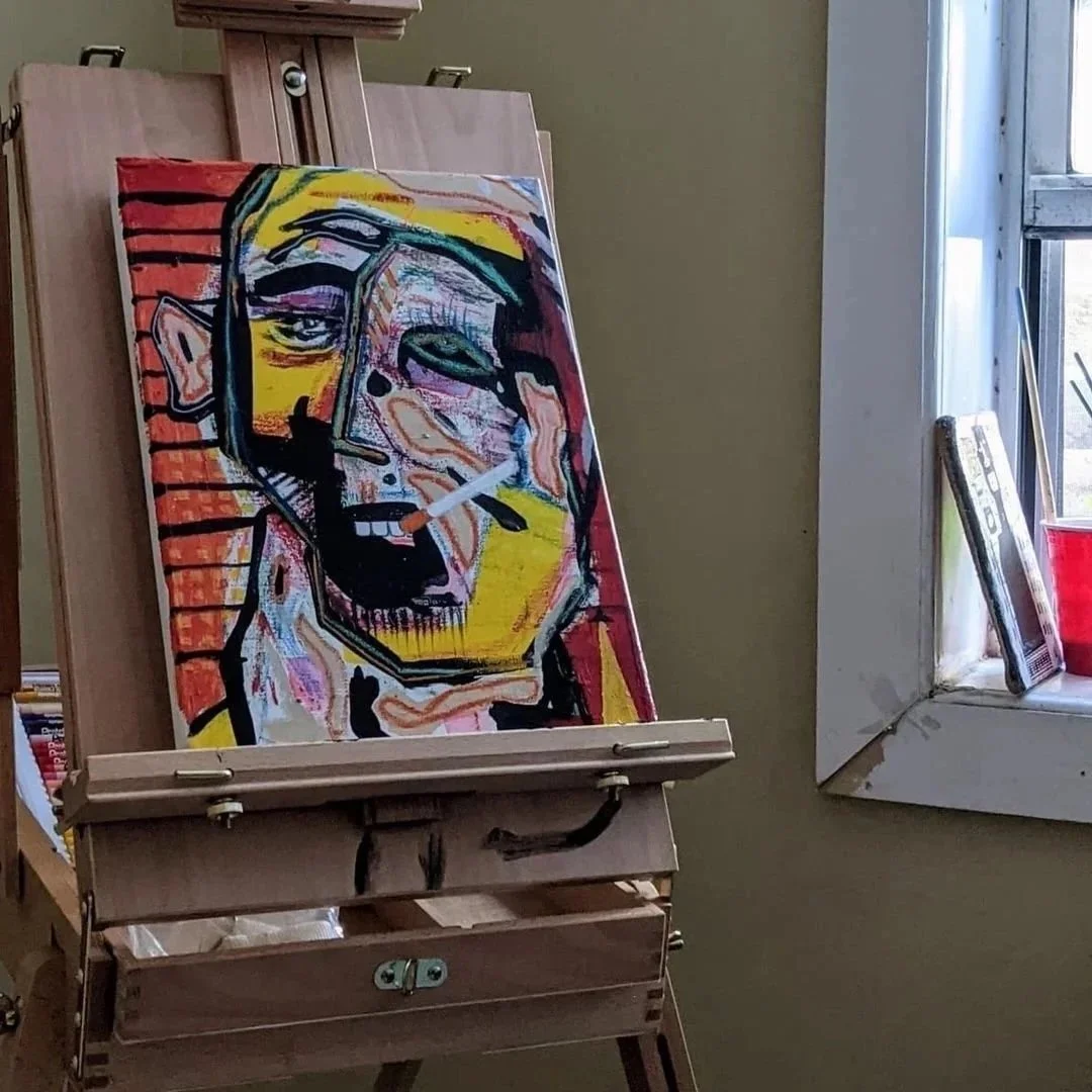A colorful abstract portrait painting on an easel inside a room near a window. The painting depicts a face with exaggerated features in bold colors like yellow, red, black, and purple.