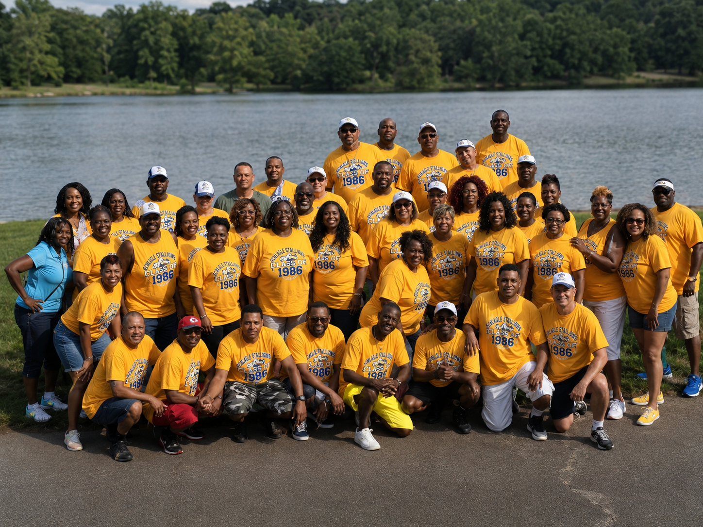 A large group of smiling people wearing yellow T-shirts with 'Class of 1986' printed on them, posing outdoors by a lake with green trees in the background.