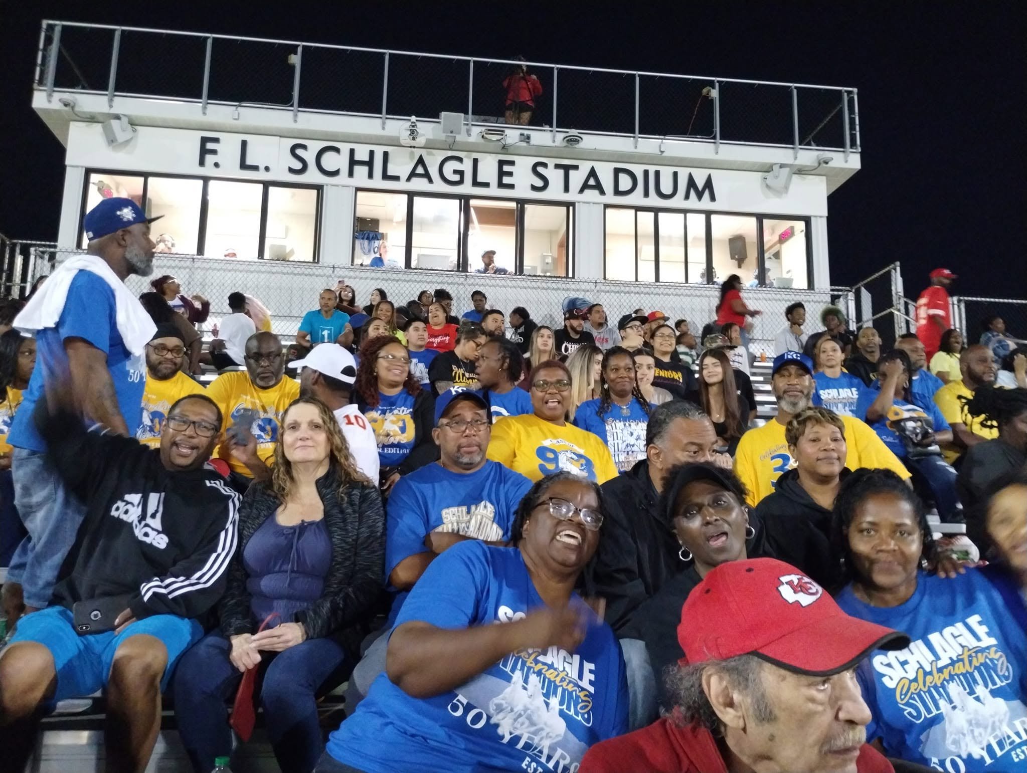 Crowd of people seated at a stadium, with a sign reading 'F. L. Schwaggle Stadium' above them. Many are wearing blue and yellow shirts, some have caps, and there is a person standing in a red shirt on the upper level.