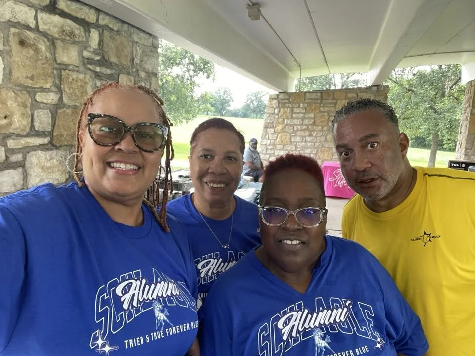 Four people smiling outdoors, wearing matching blue shirts with 'Alumni' written on them, under a pavilion with stone pillars, green trees, and grass in the background.