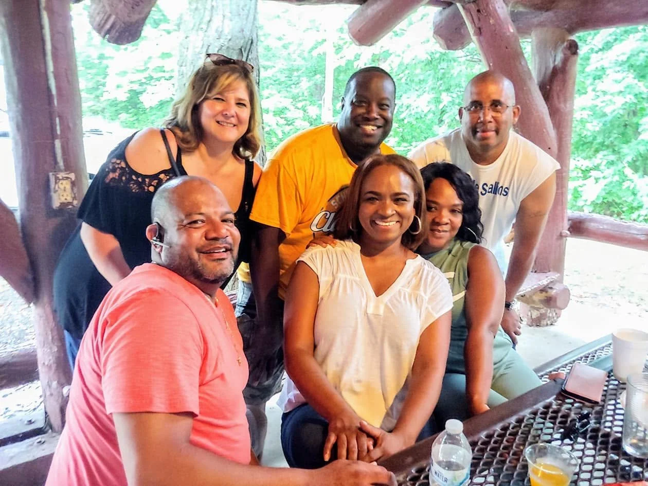 Group of six people smiling and posing around a picnic table outdoors with green trees in the background.