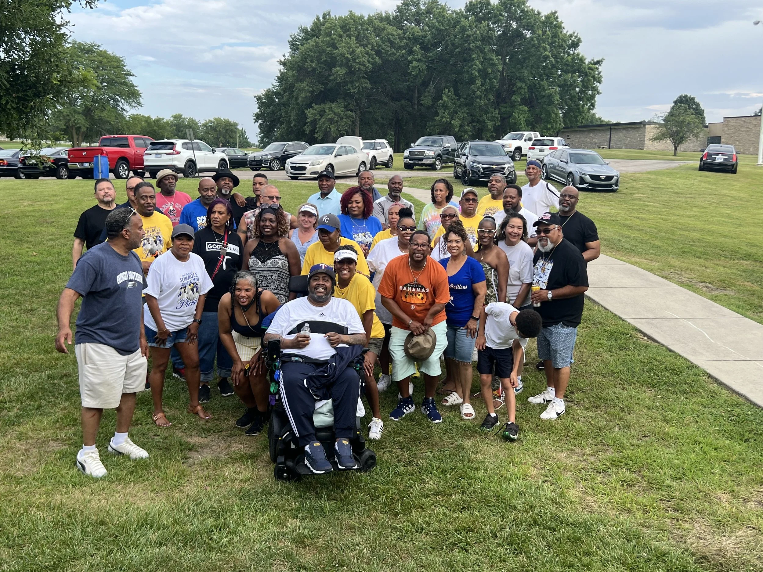 Group of diverse people posing outdoors on a cloudy day, some wearing sunglasses, standing on a grassy area with trees and parked cars in the background.