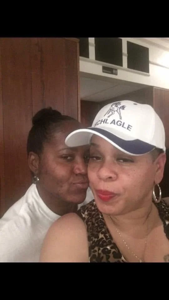 Two women taking a close-up selfie indoors. The woman on the right wears a white baseball cap with a Seahawk logo and a leopard print top, with hoop earrings and red lipstick. The woman on the left has dark skin, curly hair in a bun, and is wearing a white top with earrings. They are smiling lovingly at the camera.