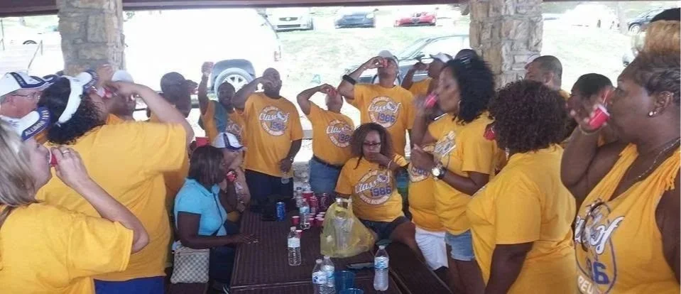 Group of women wearing yellow shirts and some with white and blue caps, standing around a table outdoors, drinking and socializing during daytime.