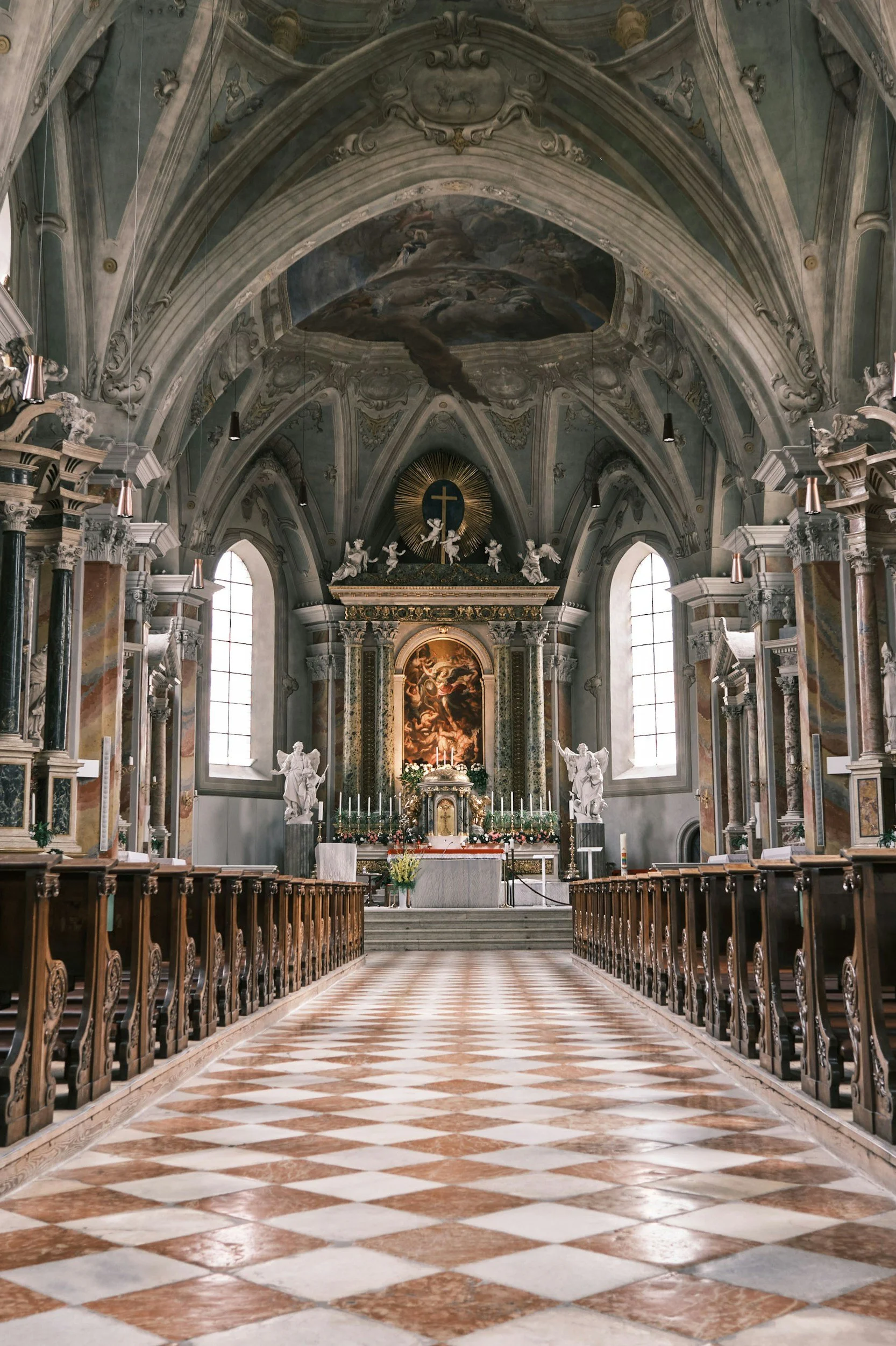 Interior of a grand church with high arched ceiling, ornate decorations, and a checkered floor leading to an altar adorned with flowers, candles, and religious artwork.
