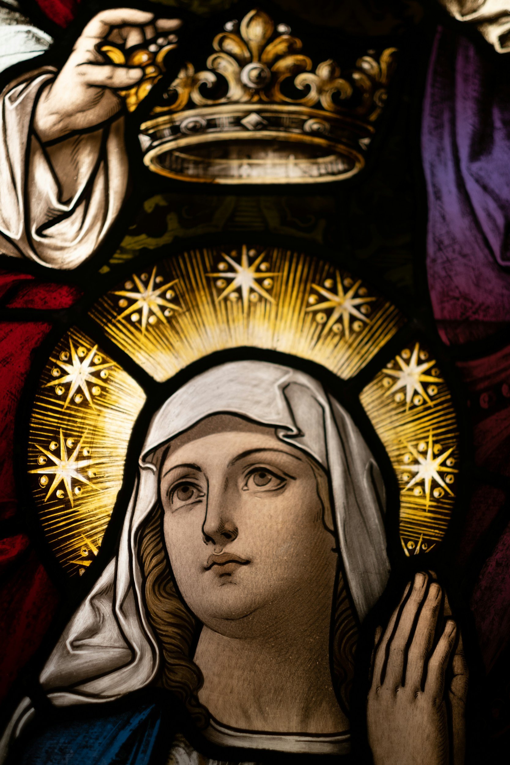 A religious stained glass window or artwork depicting the Virgin Mary, with a golden halo and surrounded by intricate golden star patterns.
