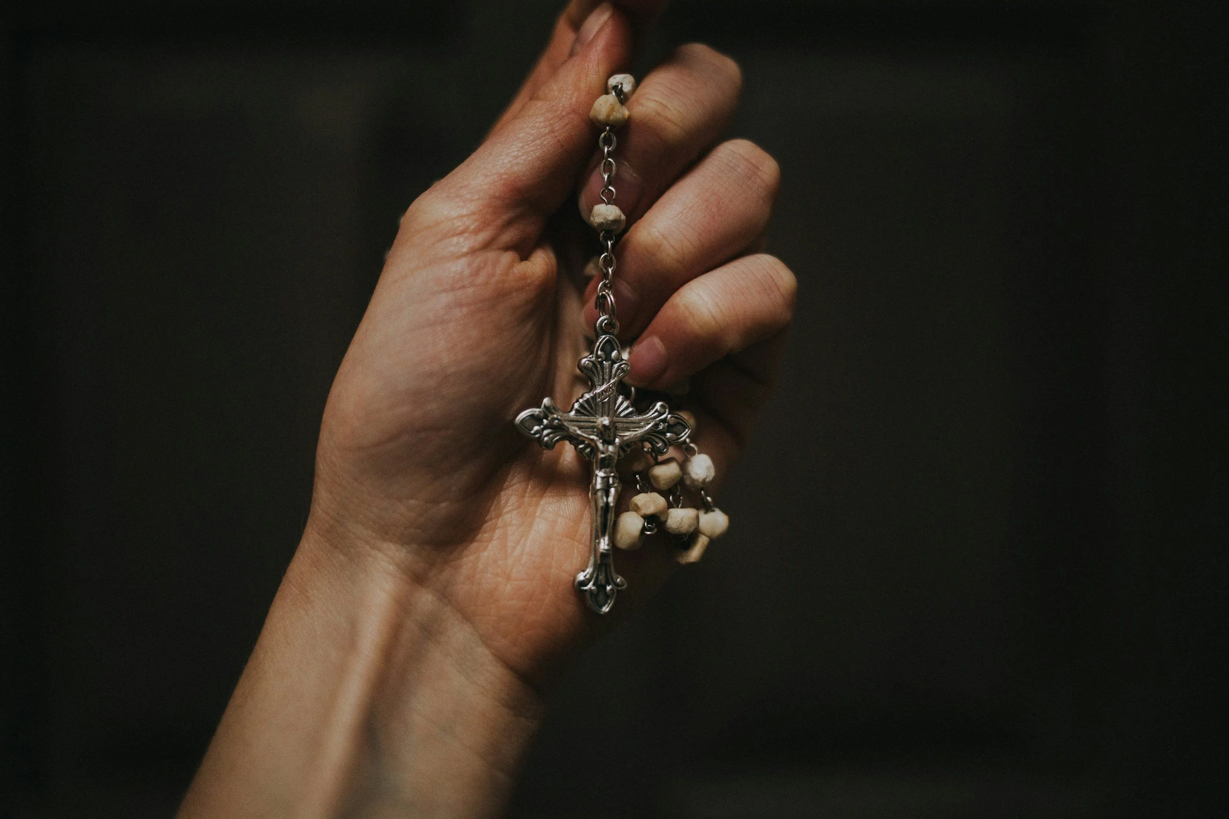 A hand holding a rosary with a metal crucifix and beads made of small stones or pearls, against a dark background.