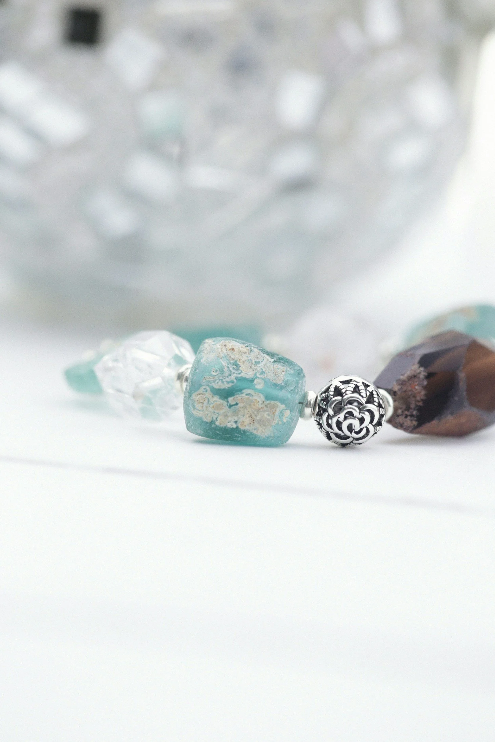 Close-up of various stones and beads, including a turquoise-colored stone, a black and white patterned bead, and other semi-precious stones, against a blurred background.