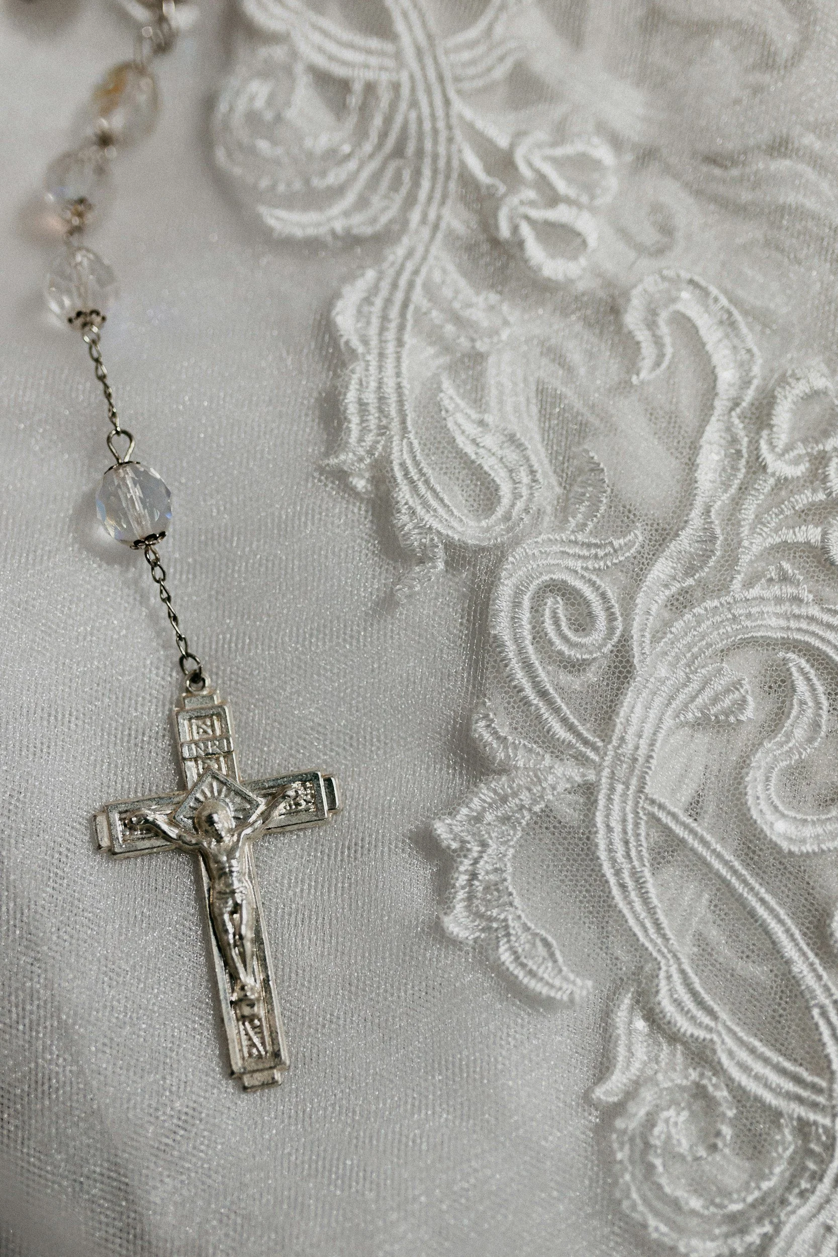 A silver crucifix necklace with a Jesus figure and a clear faceted bead, lying on white embroidered fabric with floral and swirl patterns.