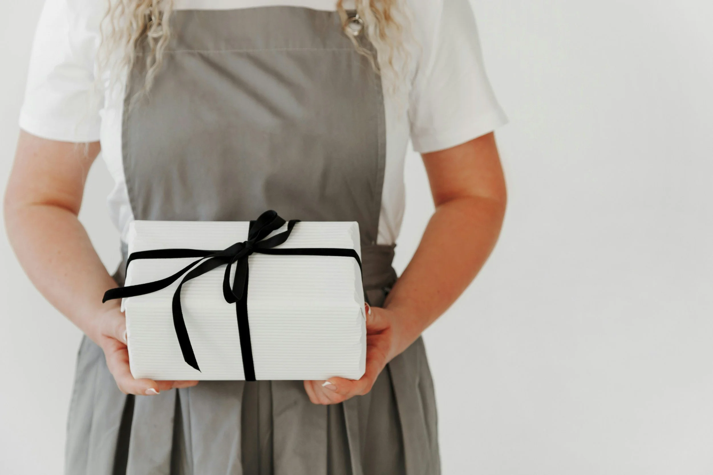 A woman wearing a gray apron over a white shirt, holding a wrapped gift box with black and white stripes and black ribbon, against a plain white background.