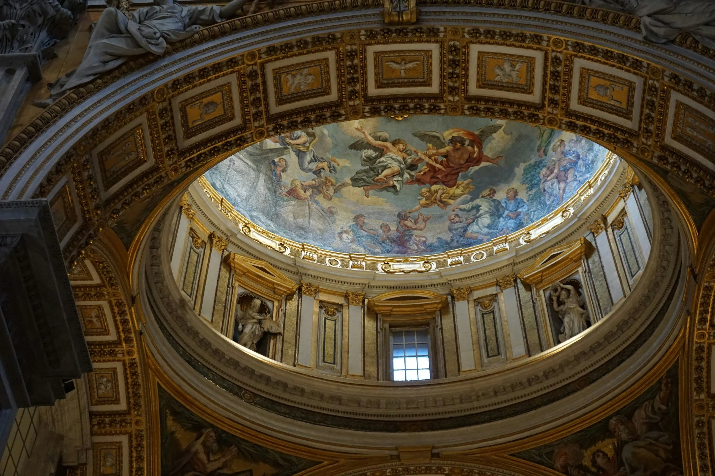 The image shows the ornate ceiling of a grand building, decorated with intricate gold details and classical paintings. The central part features a colorful mural depicting mythological figures and angels, set within a domed structure with windows allowing natural light.