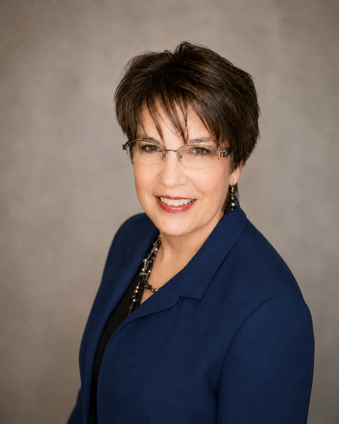 A professional woman with short brown hair wearing glasses, a blue blazer, a necklace, and earrings, smiling against a grey background.