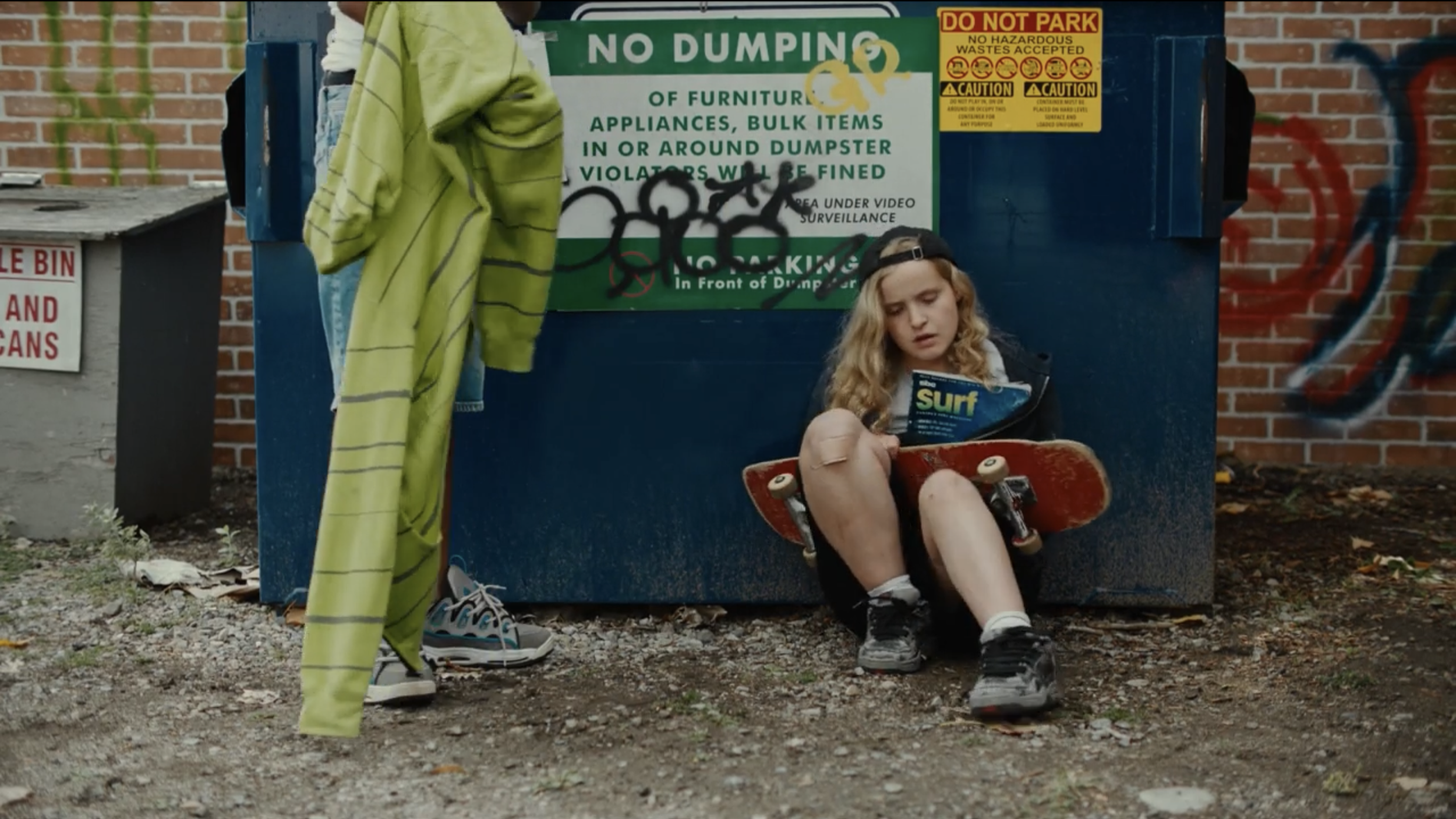 A girl with blonde curly hair sitting on the ground, holding a skateboard, and reading a magazine. Behind her, there is a blue dumpster with graffiti and signs about no dumping and no parking. Part of a person wearing sneakers and  a yellow towel is visible standing nearby, with green and yellow clothing.