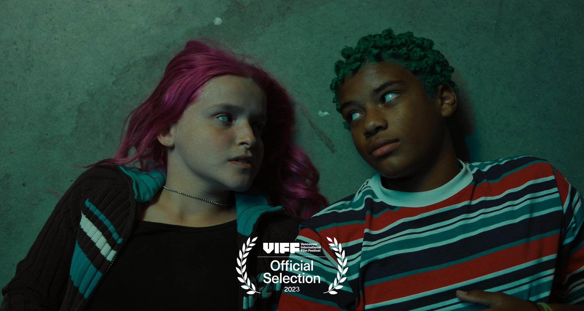 Two young women with colorful hair lying on the ground, facing each other. One has pink hair, and the other has green curly hair. They appear to be talking or sharing a moment. There is a Vancouver International Film Festival badge at the bottom of the image.