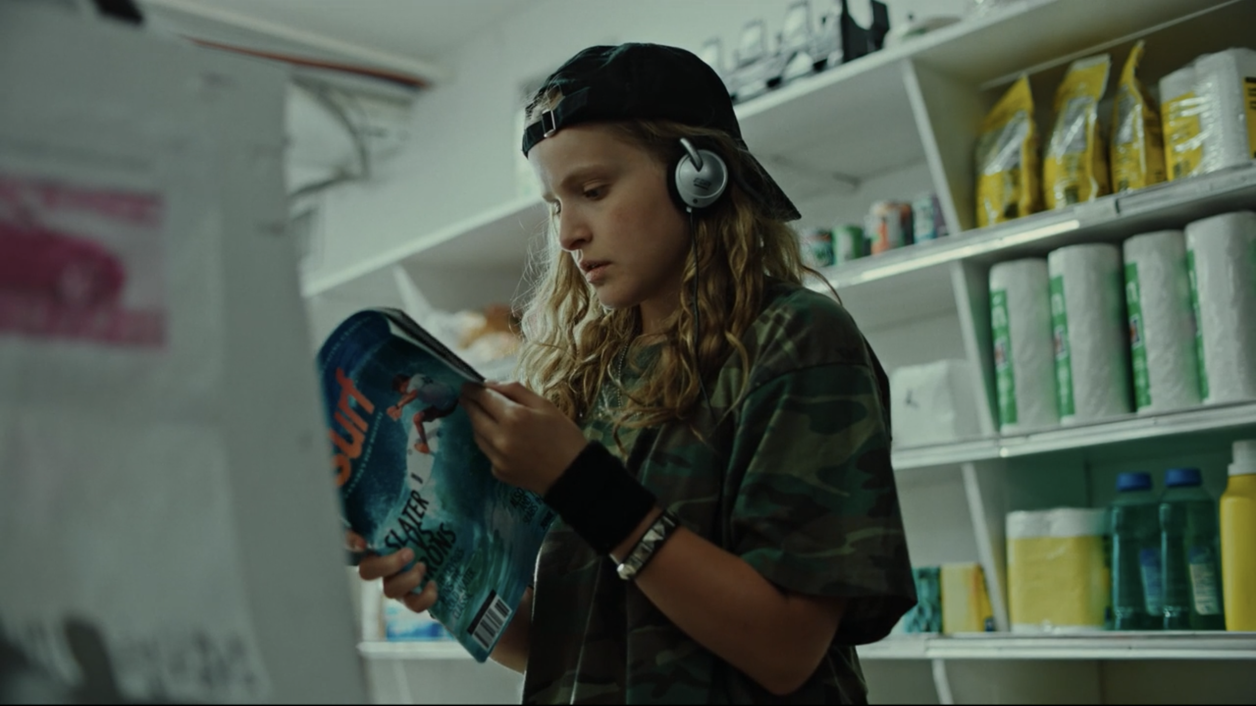 A young person with long, wavy hair, wearing a backward black baseball cap and camouflage shirt, shopping in a store aisle. They are wearing large over-ear headphones and reading a magazine or comic book.