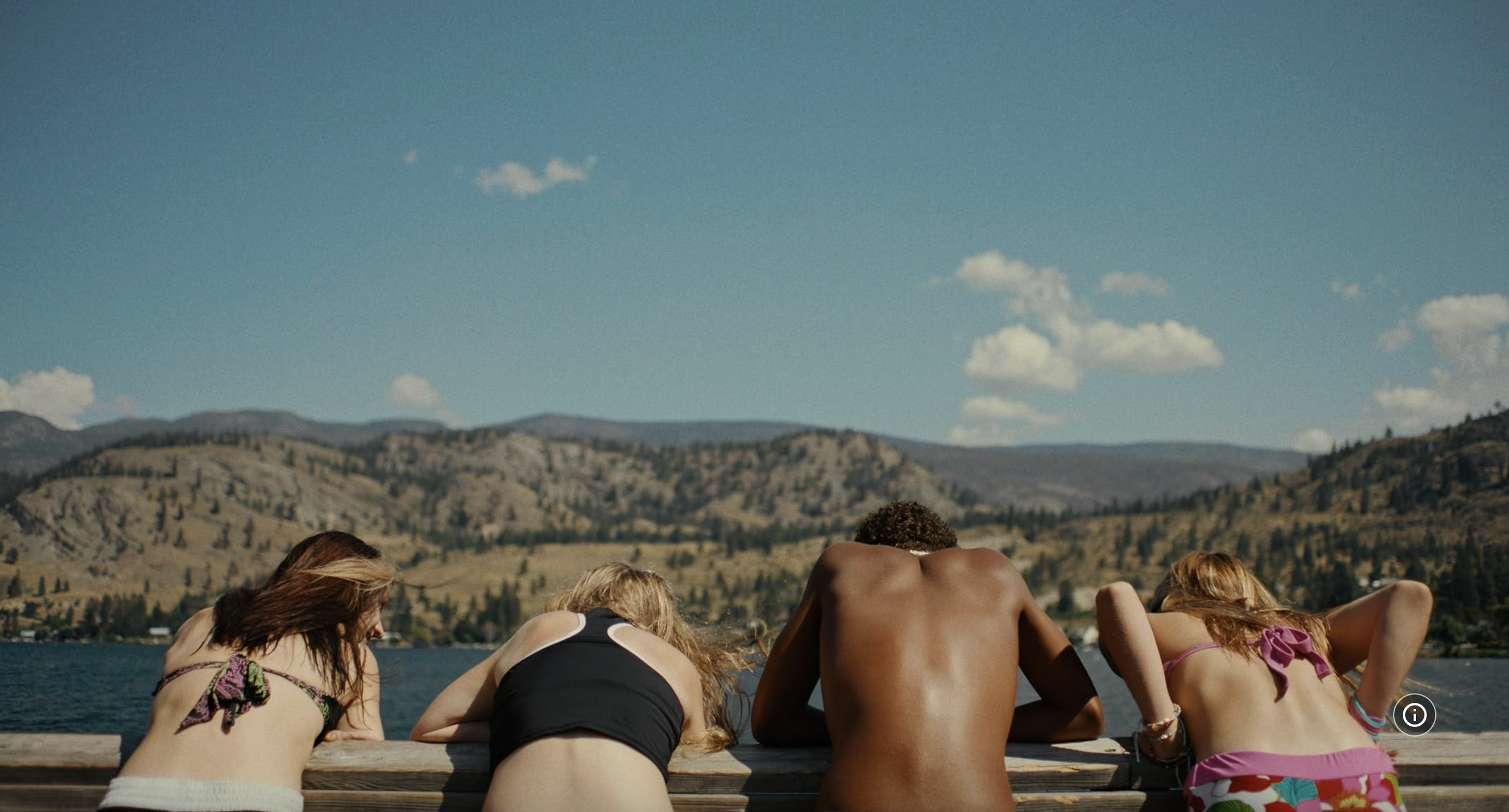 Four young people lying on a dock by a lake with mountains and blue sky in the background.