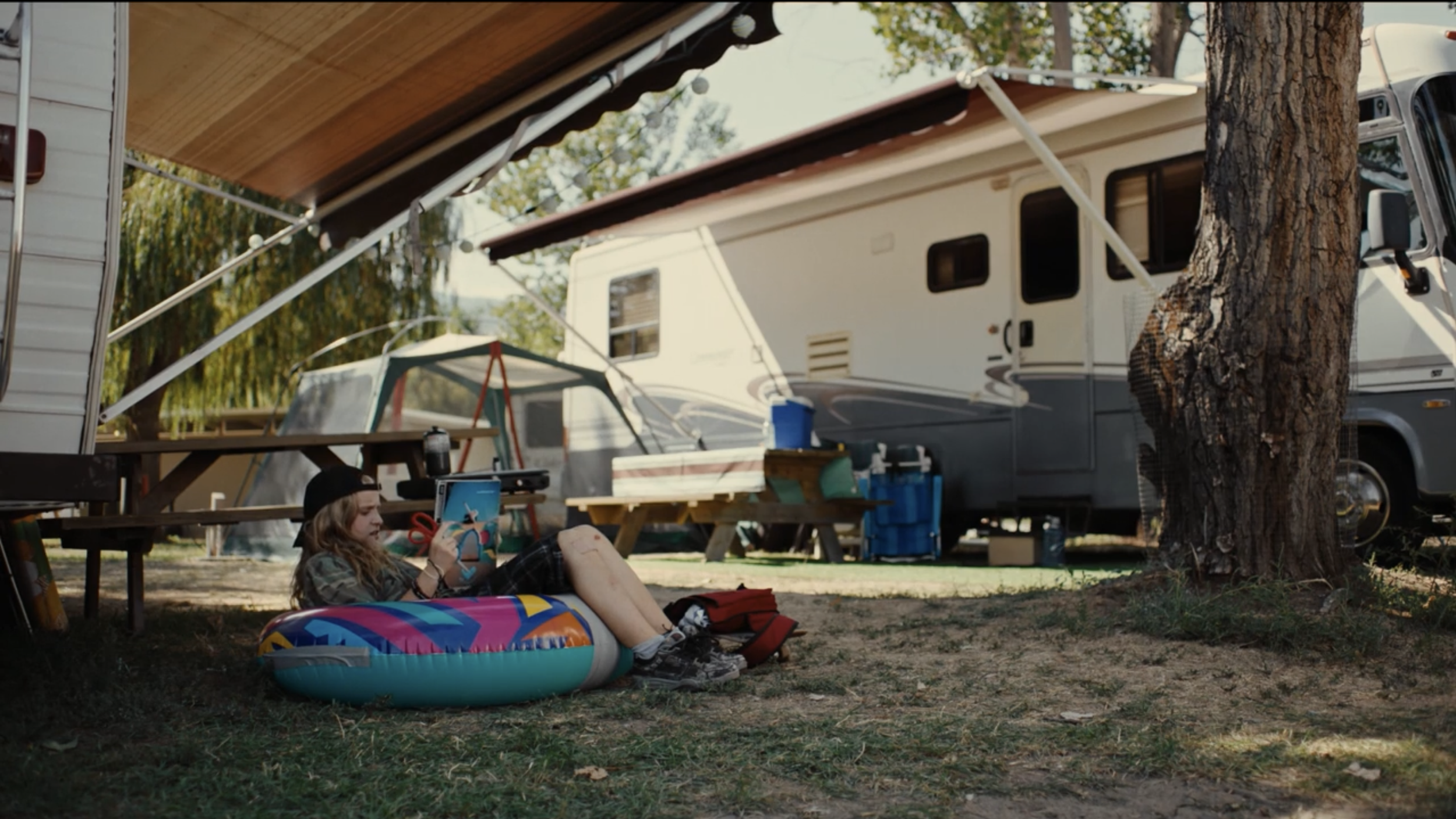 A girl relaxing on the grass in a campground with a camper in the background, surrounded by trees.