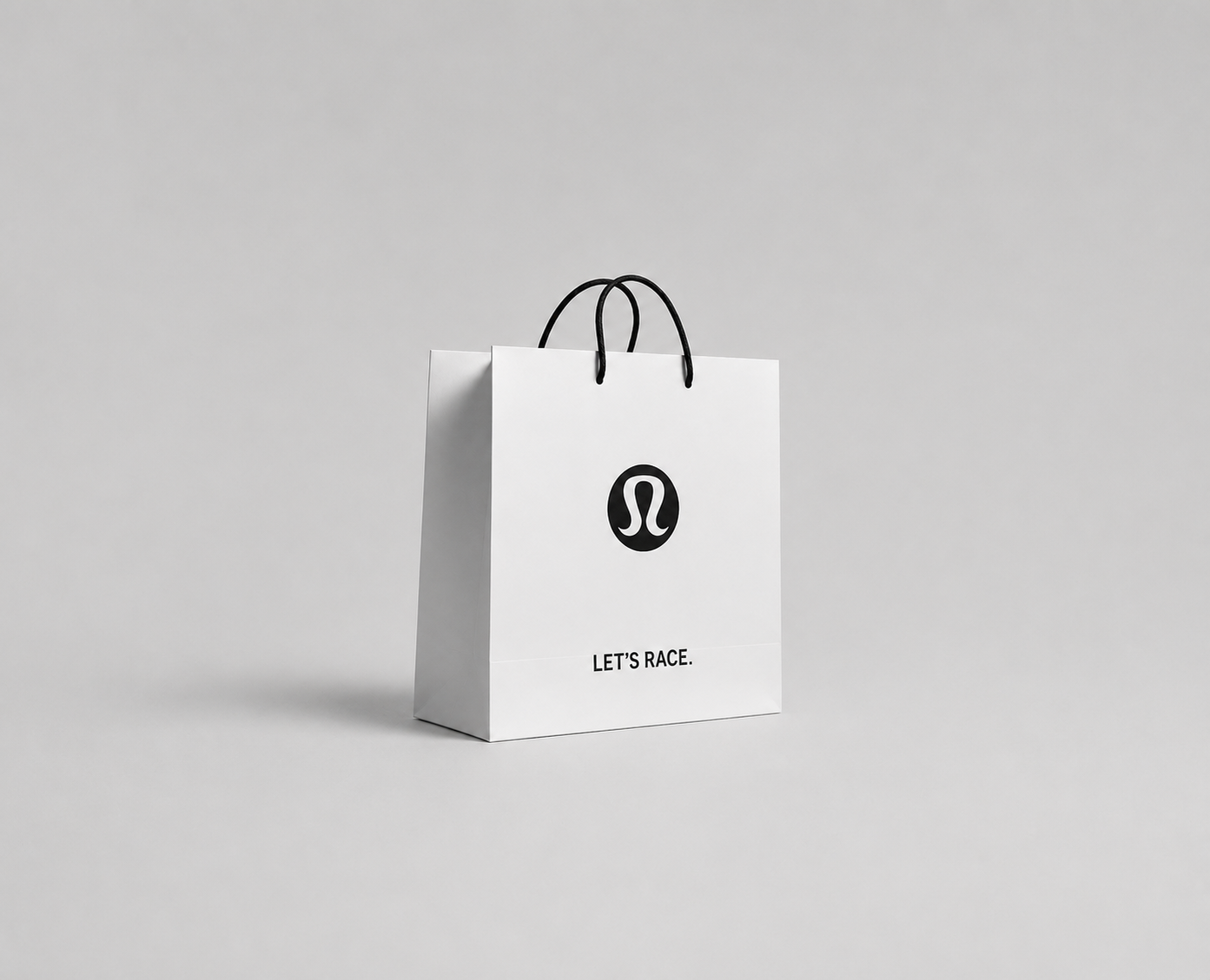 A white shopping bag with black handles and logo, with the phrase "LET'S RACE" printed on it, on a plain light grey background.