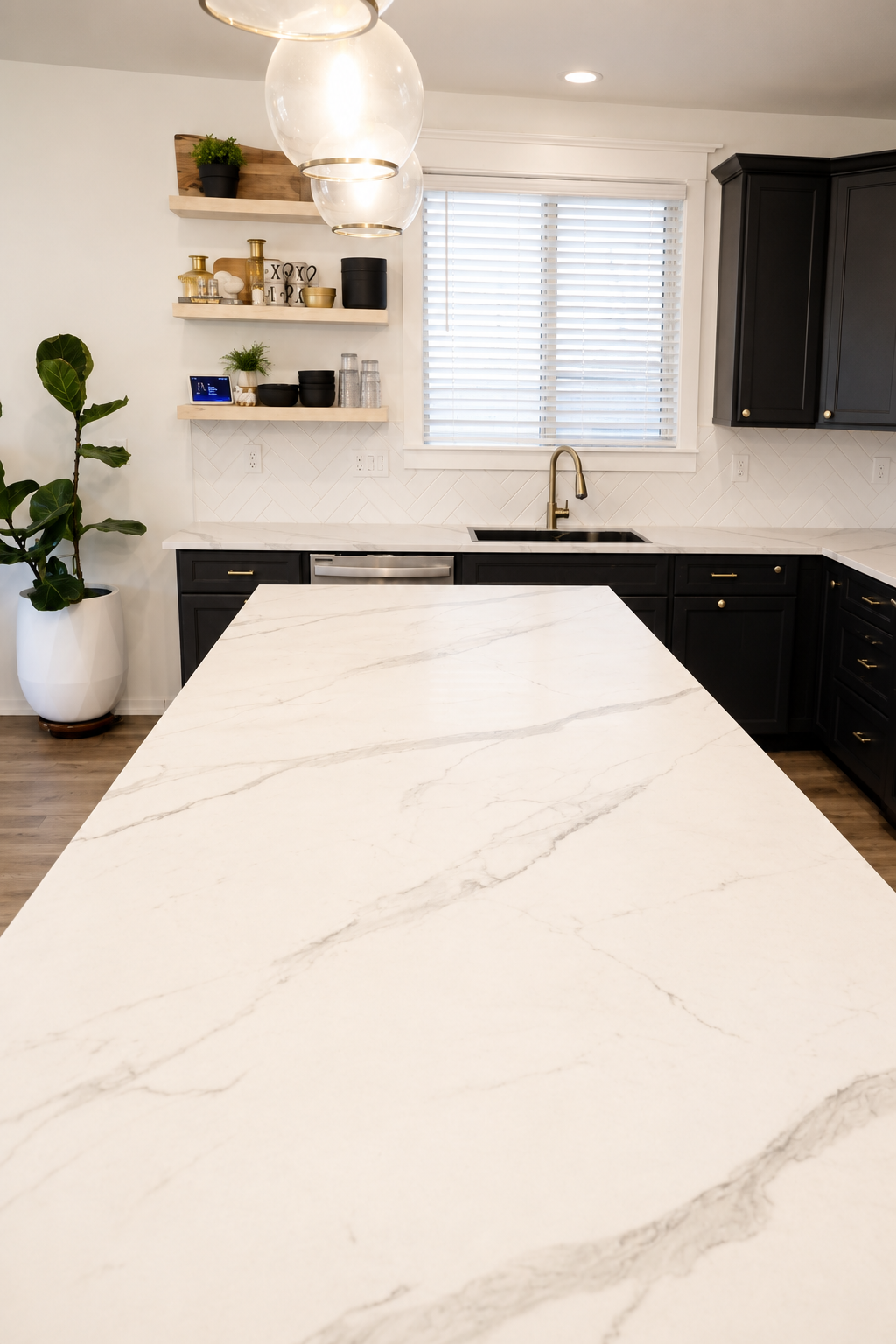 Modern kitchen with a white marble island, black cabinets, gold hardware, and a window with white blinds. Decor includes potted plants and open wooden shelves with black, gold, and white dishes and accessories. ReSlabb method to cover and refinish st