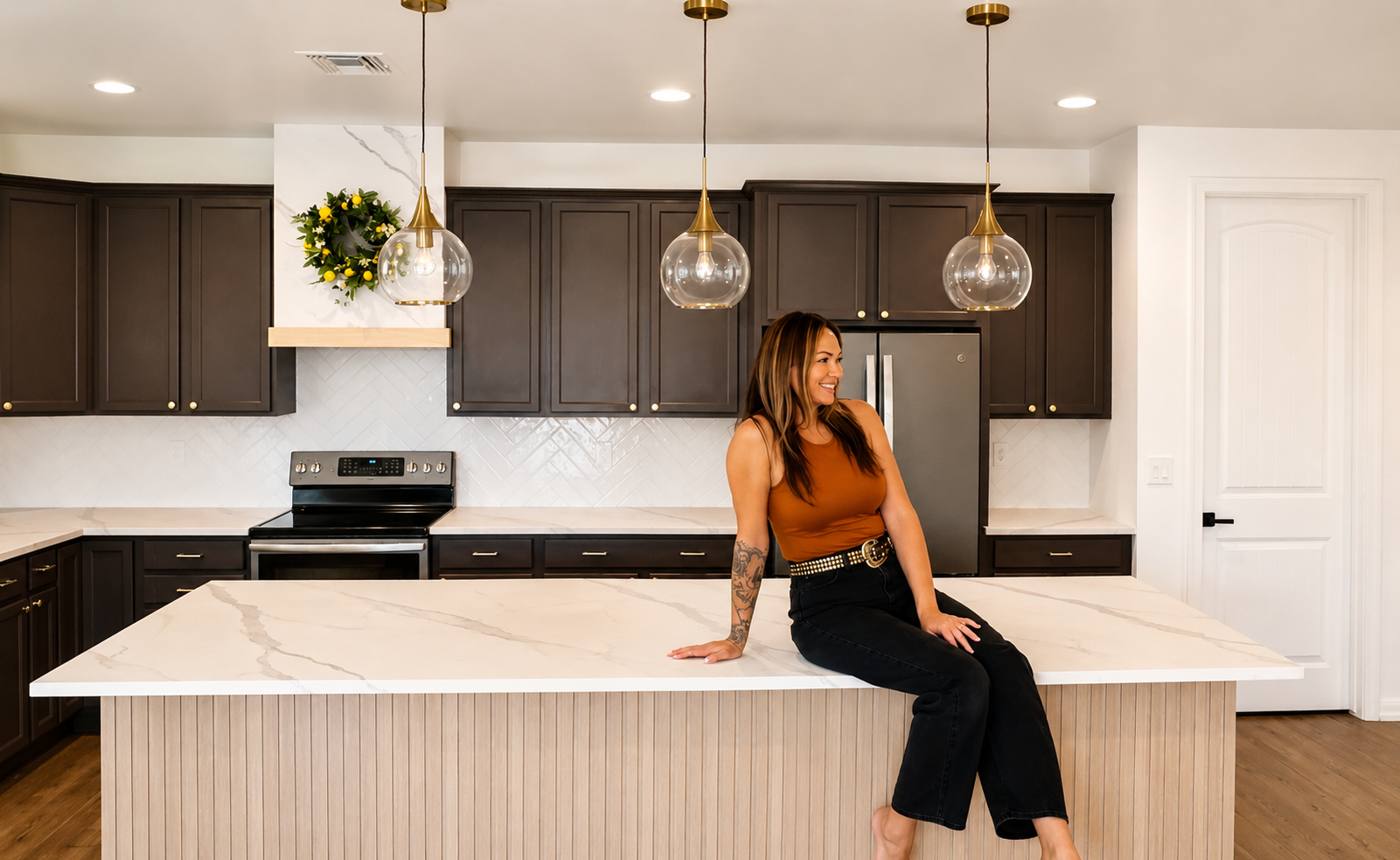 A woman with long brown hair, wearing a rust-colored sleeveless top and black jeans with a studded belt, sitting on a kitchen island with a white marble countertop. The kitchen has dark brown cabinets, stainless steel appliances, pendant lights, and a decorative lemon and greenery wreath on the wall.
