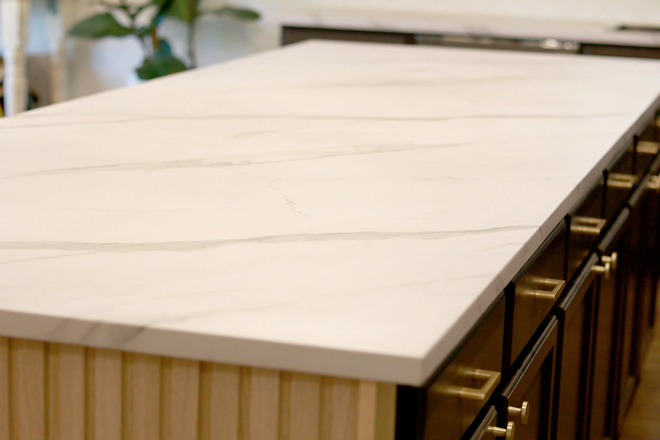 ReSlabb method to cover and refinish stone countertops. A large white marble-topped island in a kitchen with light wood paneling on the sides and black cabinets with gold handles underneath.
