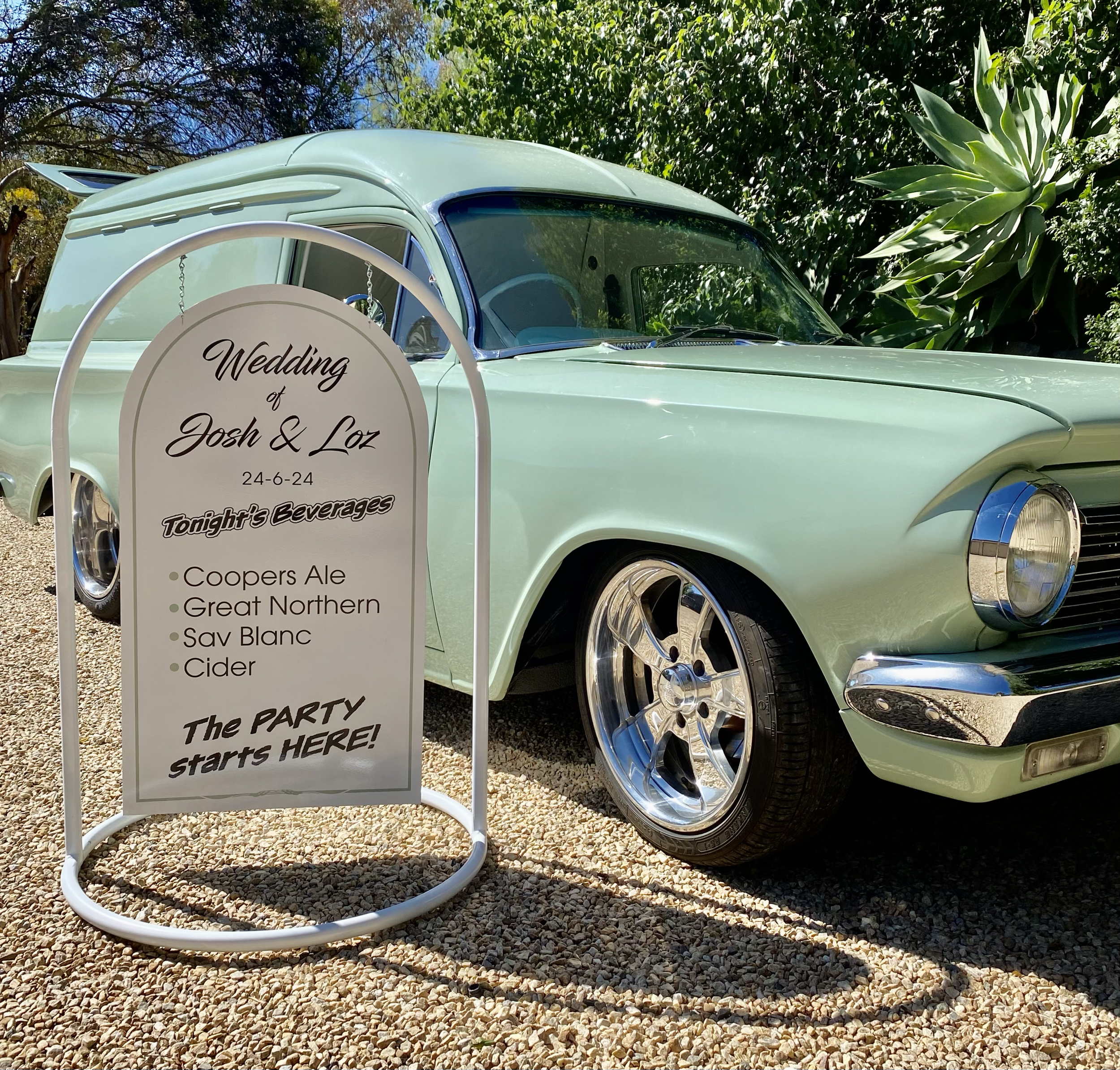 A vintage mint green car with a white circular wedding sign attached, displaying the names Josh and Loz, and an event date of June 24, 2024, parked on gravel with trees in the background.