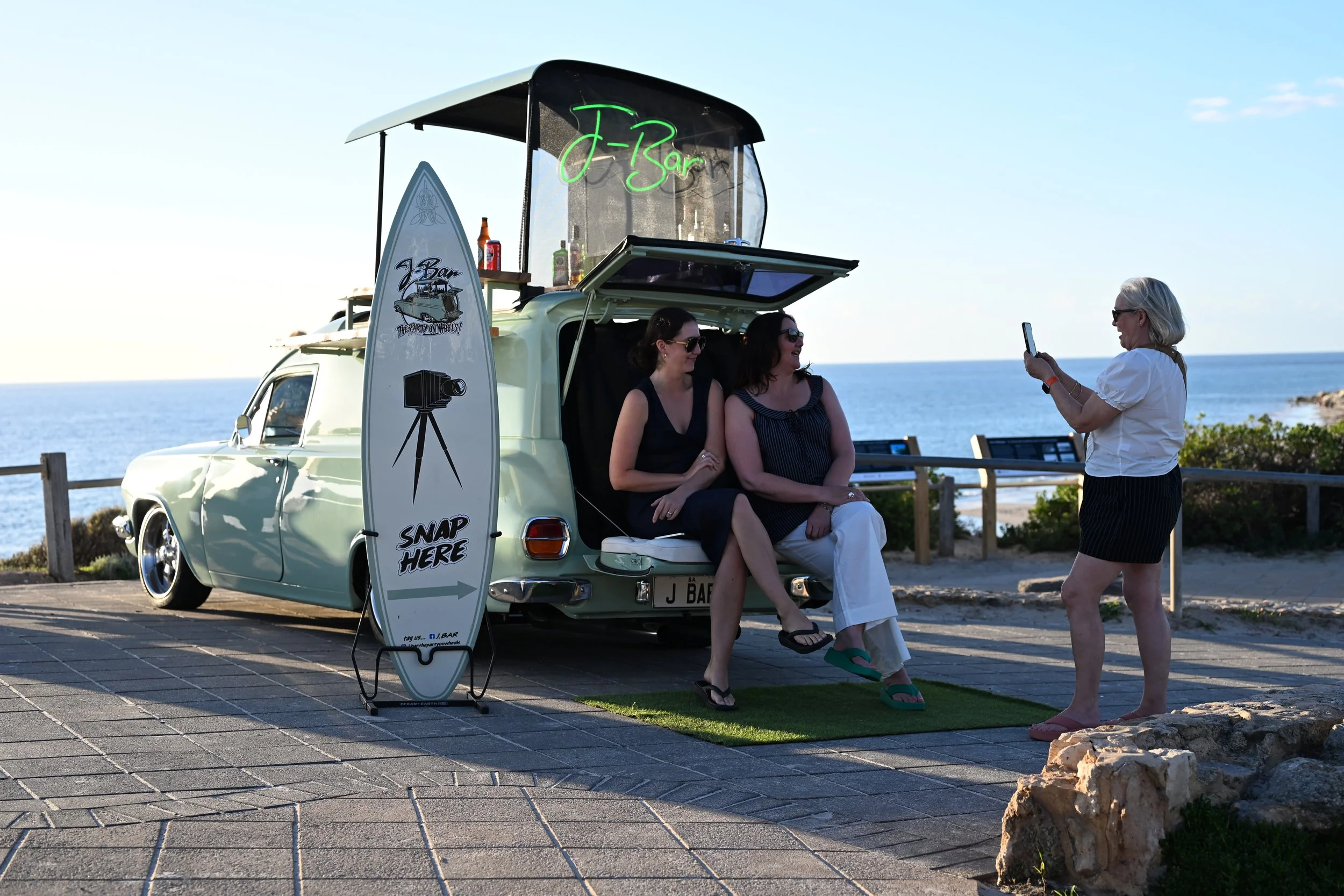 A surfboard with a sign saying 'Snap Here' and an arrow pointing to a vintage van with a beverage stand on top, where two women are seated and a third woman is taking a photo near the ocean.