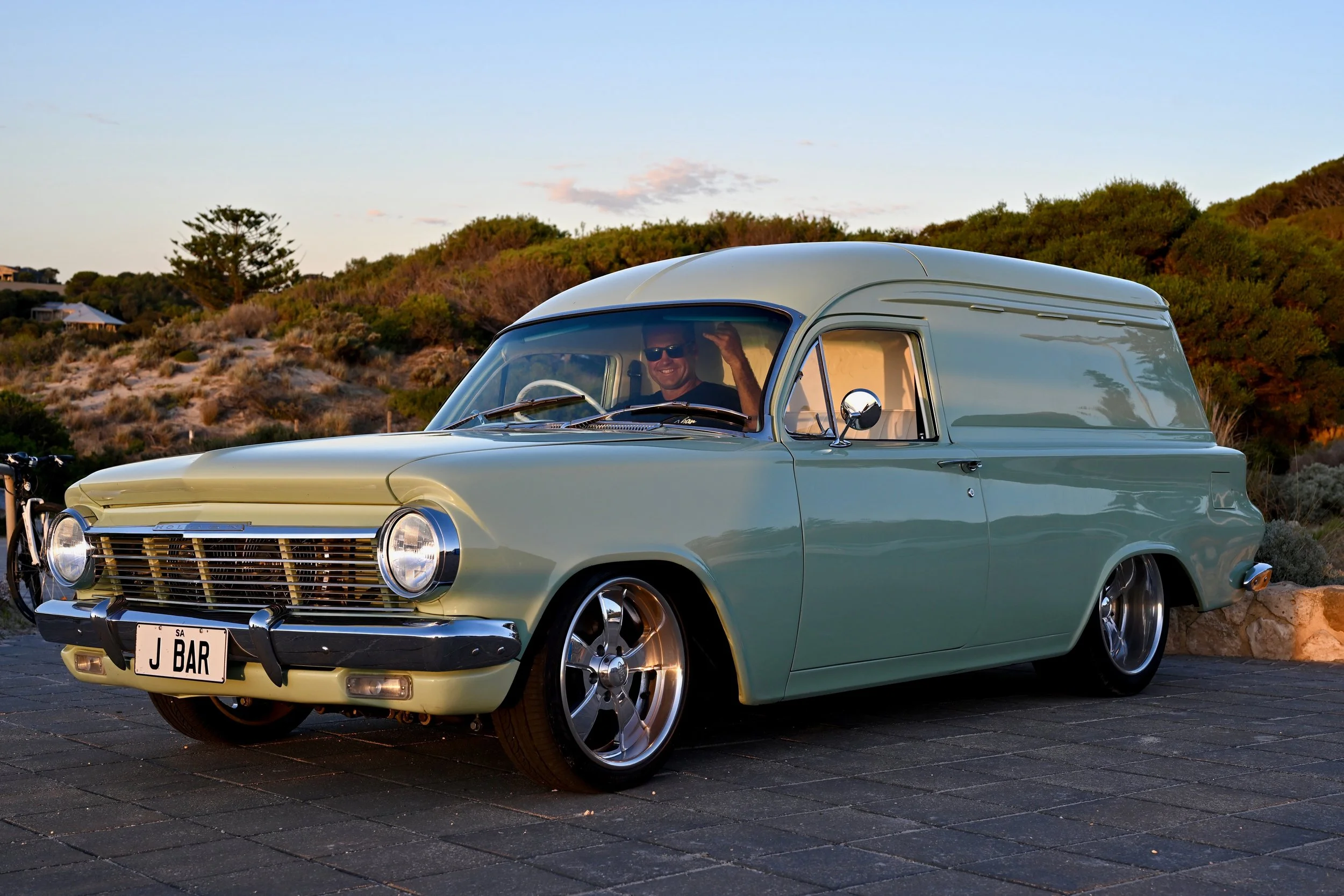 A vintage light green car with custom wheels, smiling man inside giving a thumbs-up, parked outdoors with desert and hills in the background during sunset.