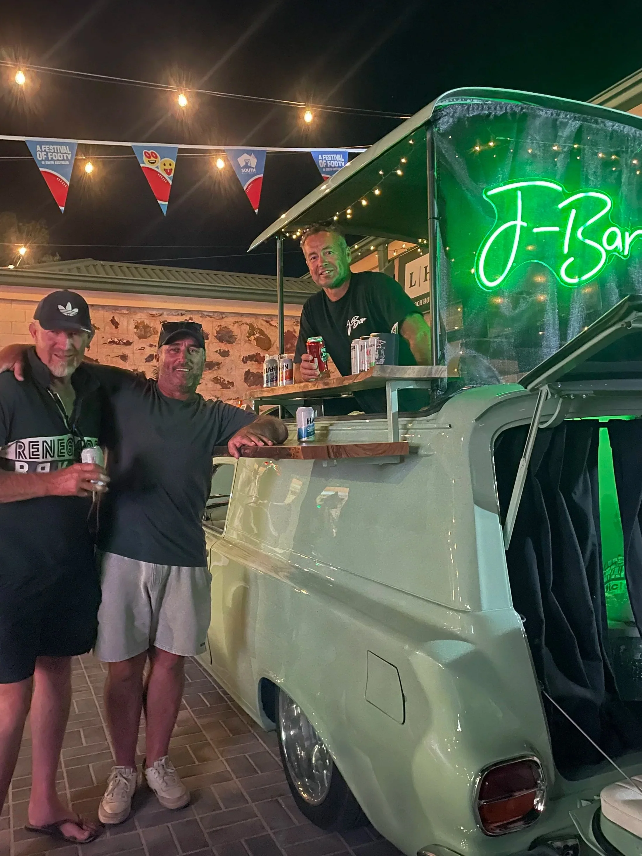 Three people standing next to a vintage green car with a J-Bar sign, at night with string lights and festival banners overhead.