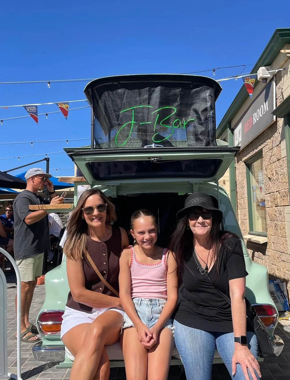Three women and one young girl sitting on the trunk of a light green vintage car with an open trunk that has a sign reading 'J Bar' in neon green. The women are smiling, and the girl is smiling, all are casually dressed in summer clothes and sunglass