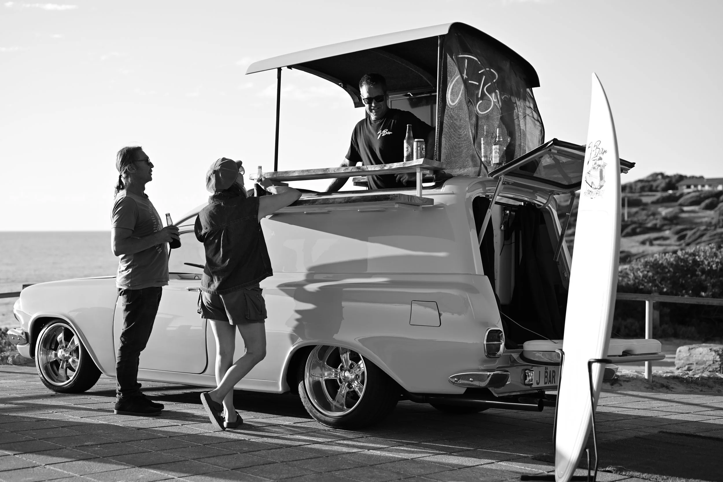 Three people near a vintage car by the beach; one person is inside a food or drink stand built on top of the car, serving two women, with the ocean visible in the background.