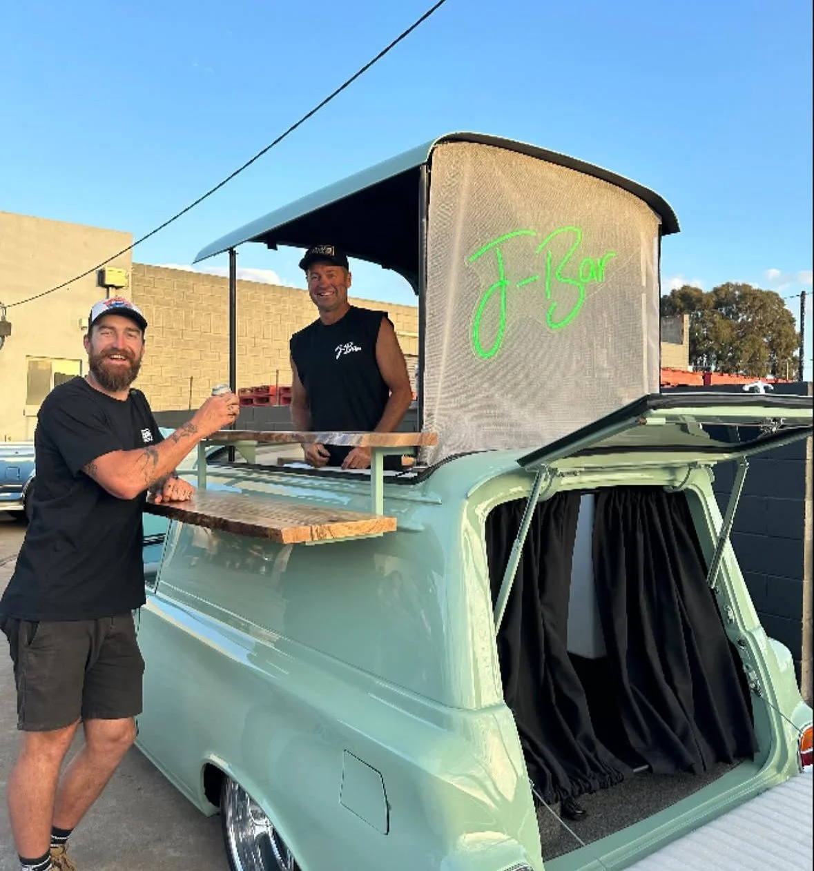 A vintage green vehicle converted into a small food stand with a sign that says 'J-Bar.' Two smiling men, one with a beard and cap, the other standing behind the counter, are serving and enjoying drinks outside on a sunny day.