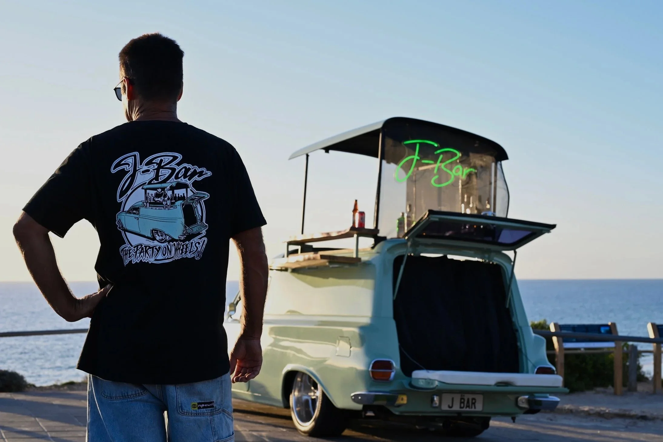 A man standing with his back to the camera, wearing sunglasses, a black T-shirt with a car graphic that says 'J-Bar' and 'The Party on Wheels', near a vintage light green car with an open trunk and a white canopy labeled 'J Bar' in stylized green neo