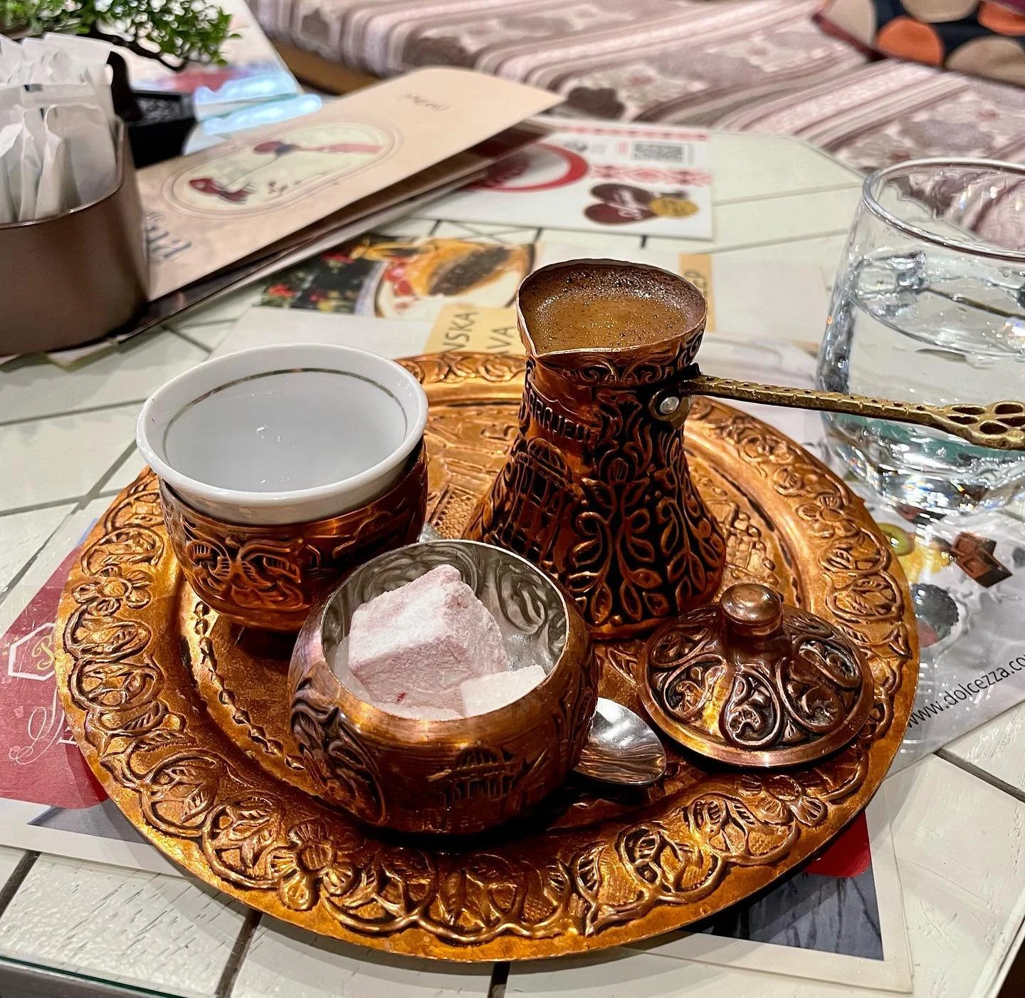 A traditional Middle Eastern coffee set on a decorative gold tray, including a cezve (coffee pot), a cup, a small container with sugar cubes, and a glass of water, all on a white tiled table with menus and a glass of water in the background.