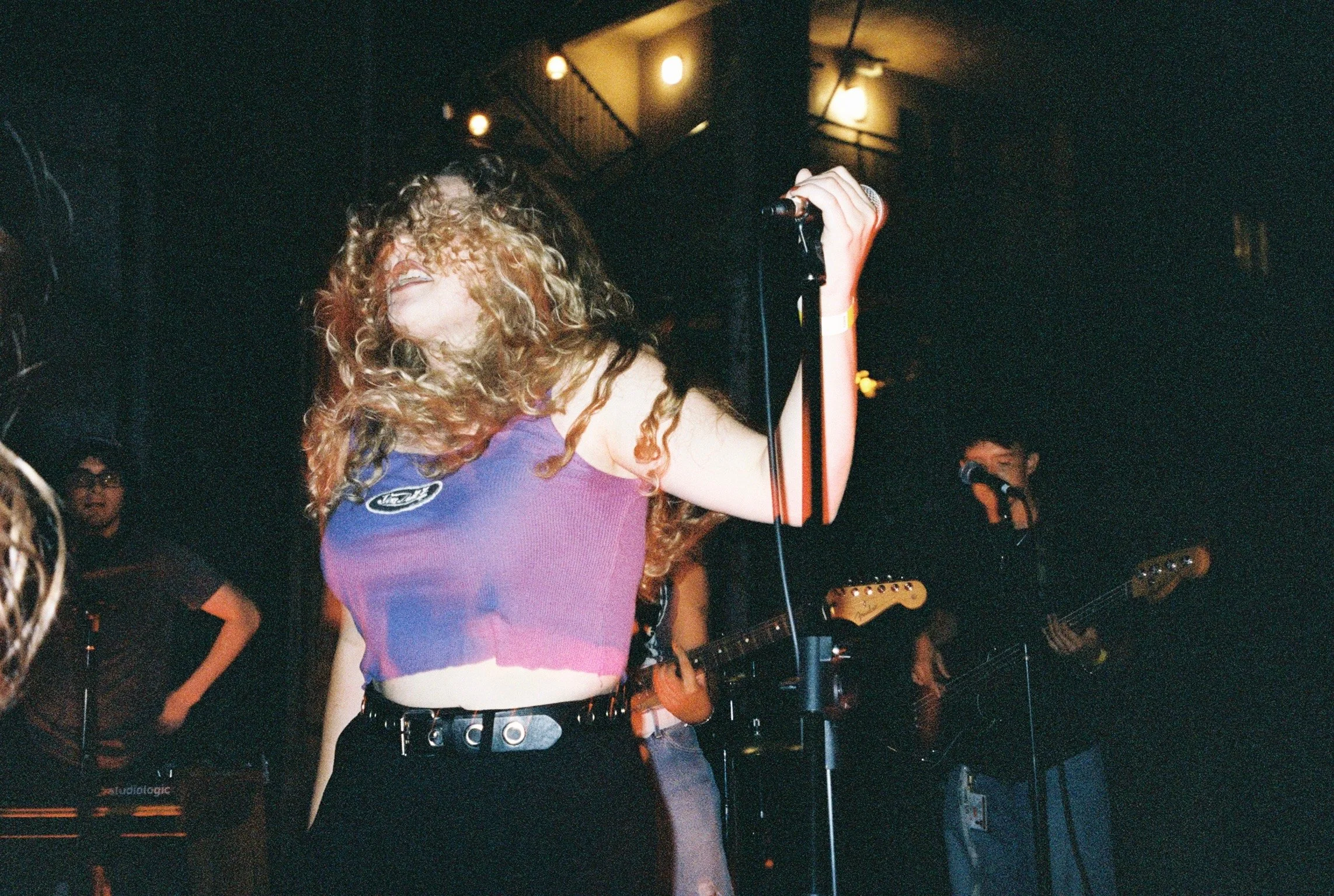 A woman with curly hair singing into a microphone on stage, wearing a purple top, with band members and music equipment in the background.